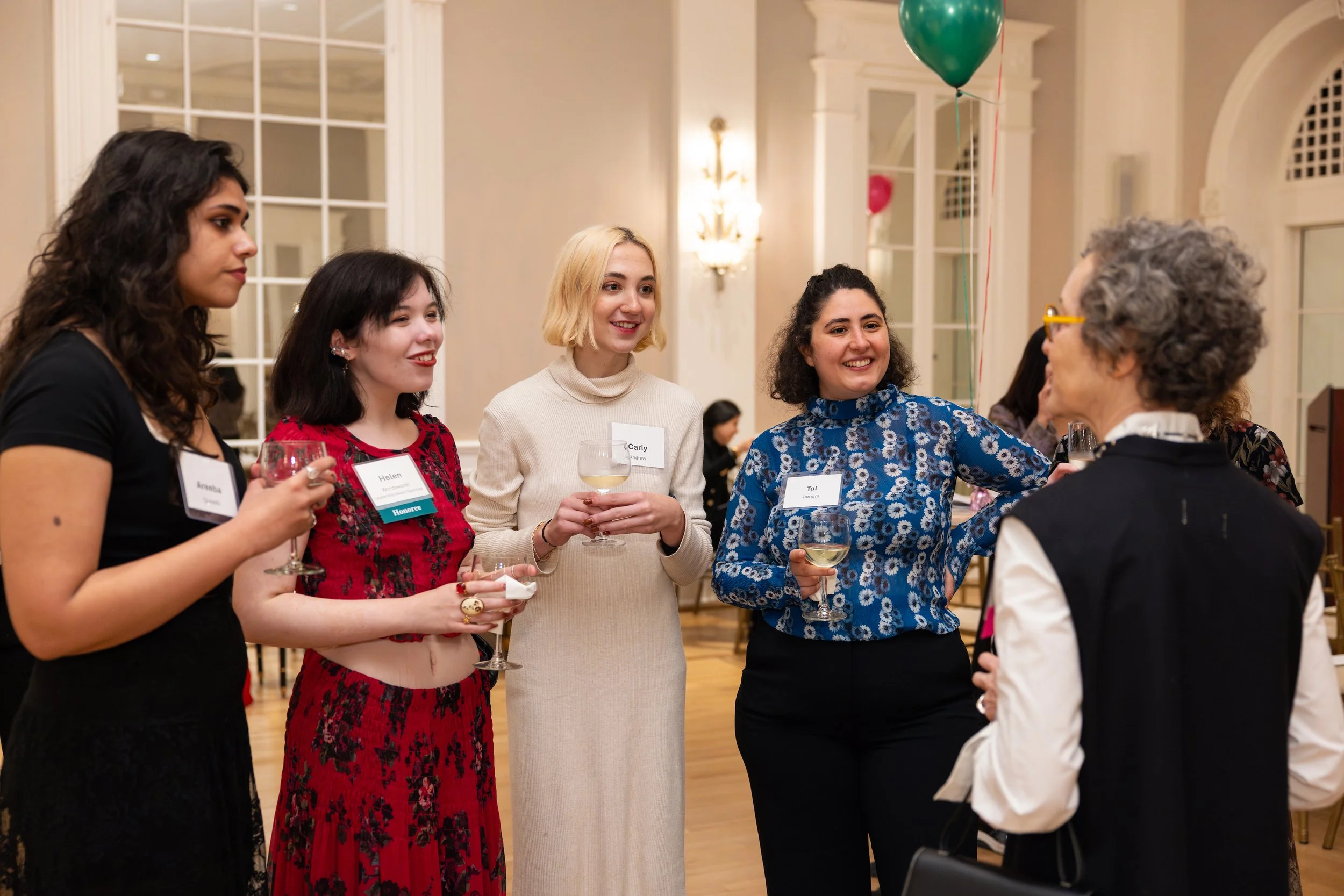 A group of five women at a social event, holding glasses of wine and engaging in conversation. They are in an elegant room with high windows and warm lighting, wearing name tags.