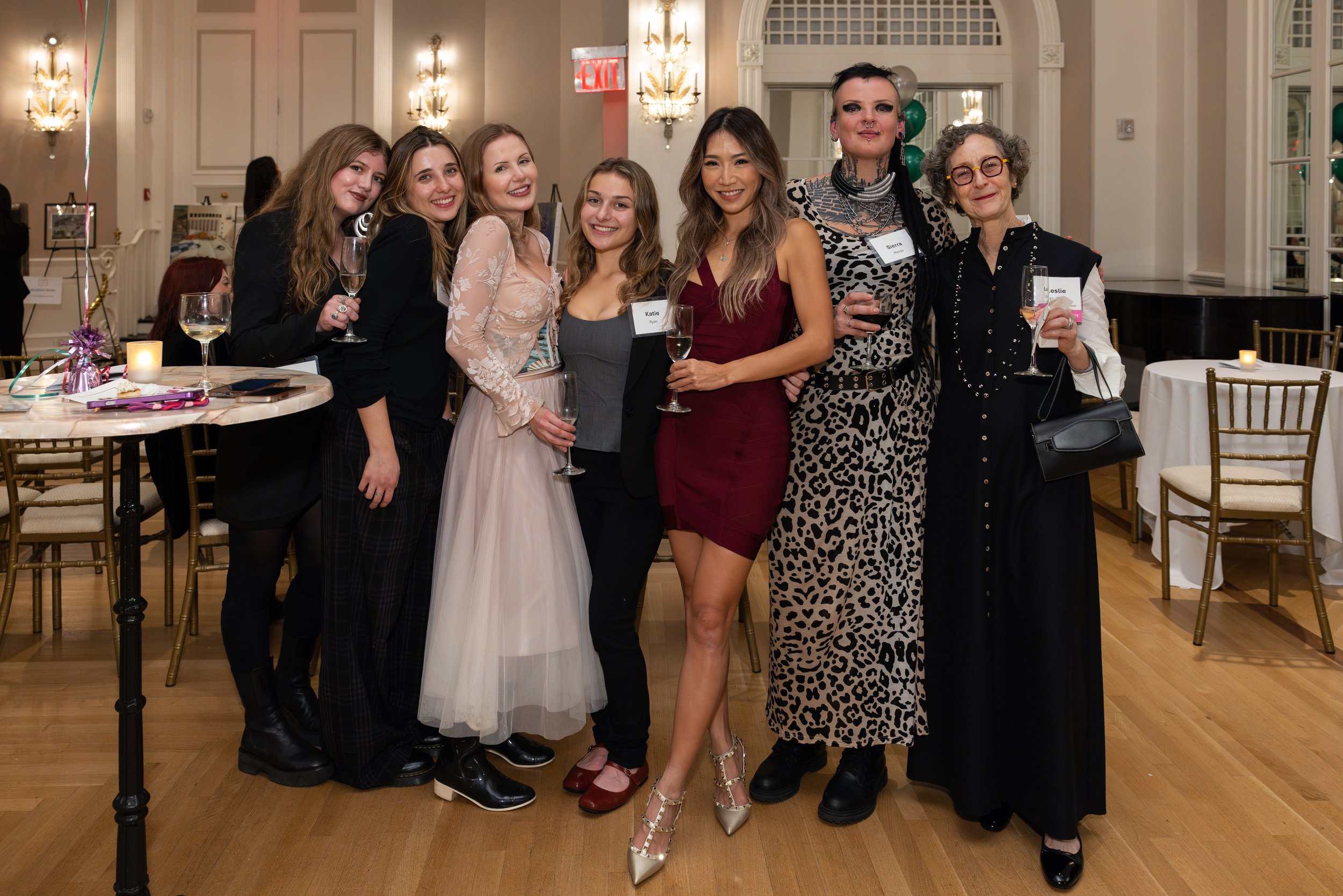 Group of eight women standing together at a celebration, smiling and holding wine glasses, in an elegant indoor setting with chandeliers and decorated tables.