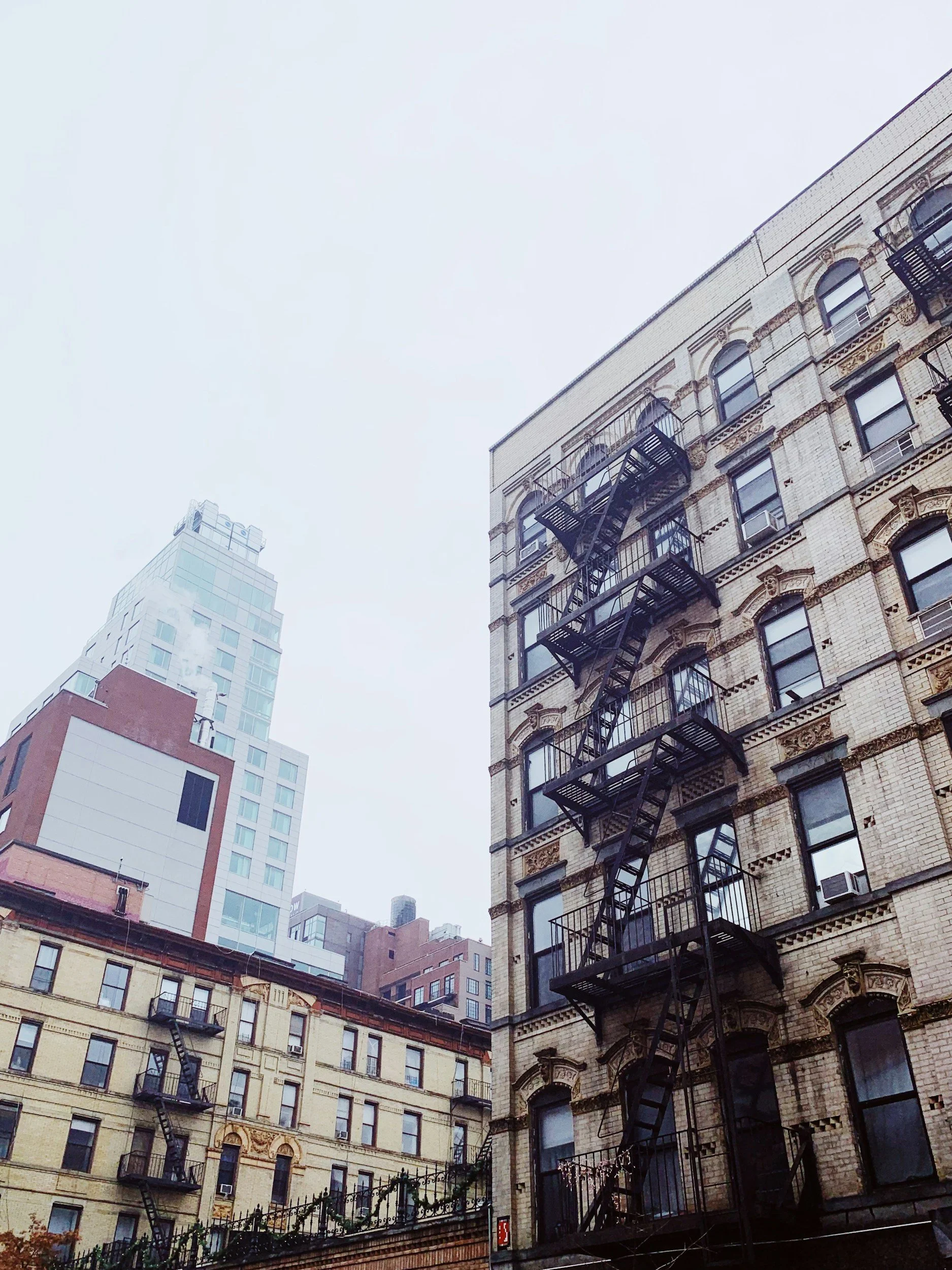 Tall city buildings with fire escapes on the exterior of the buildings, viewed from the ground looking up on a cloudy day.