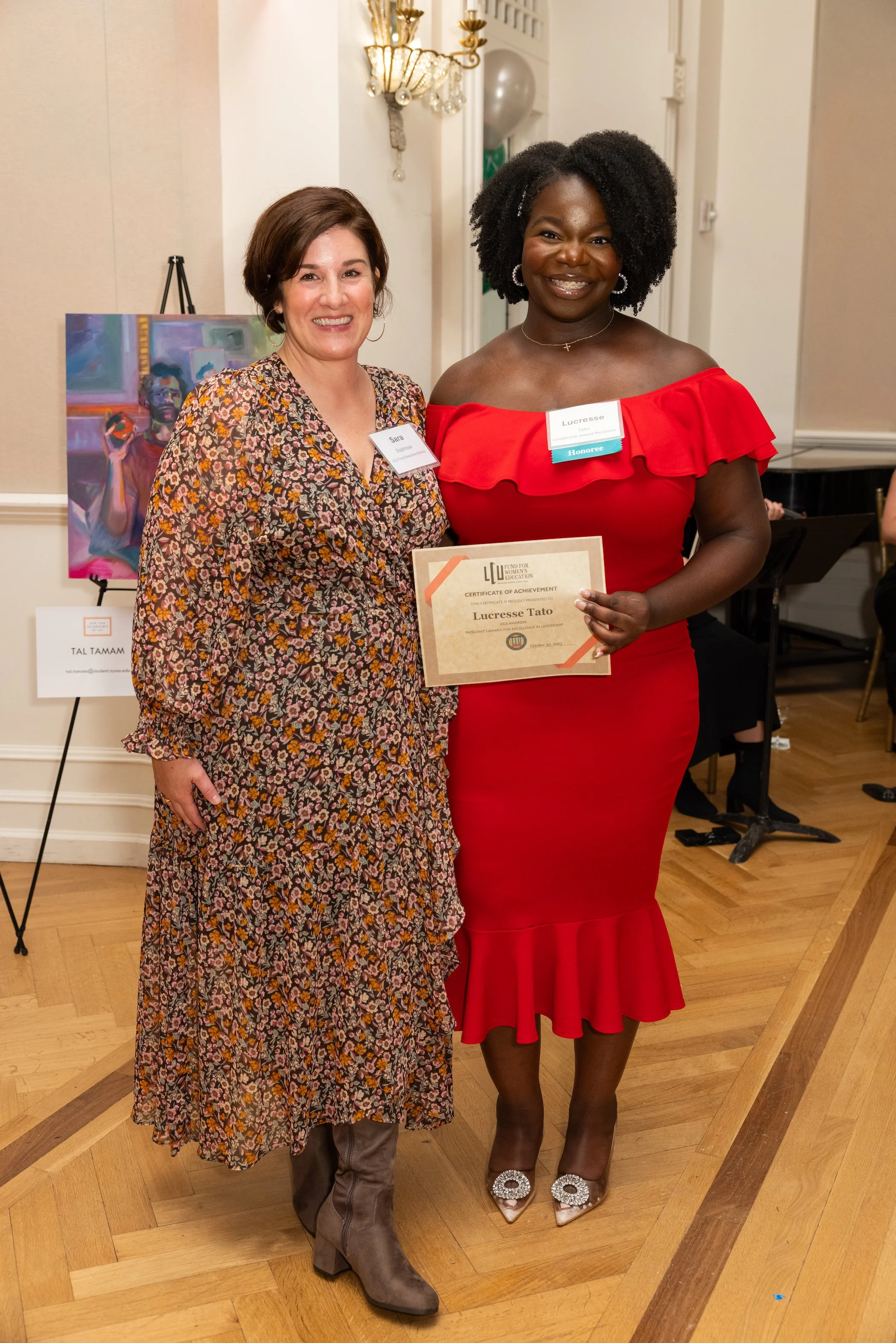 Two women standing together at an award ceremony, one holding a certificate. The woman on the left has light skin, short brown hair, and is wearing a floral dress with brown boots. The woman on the right has dark skin, curly black hair, and is dresse