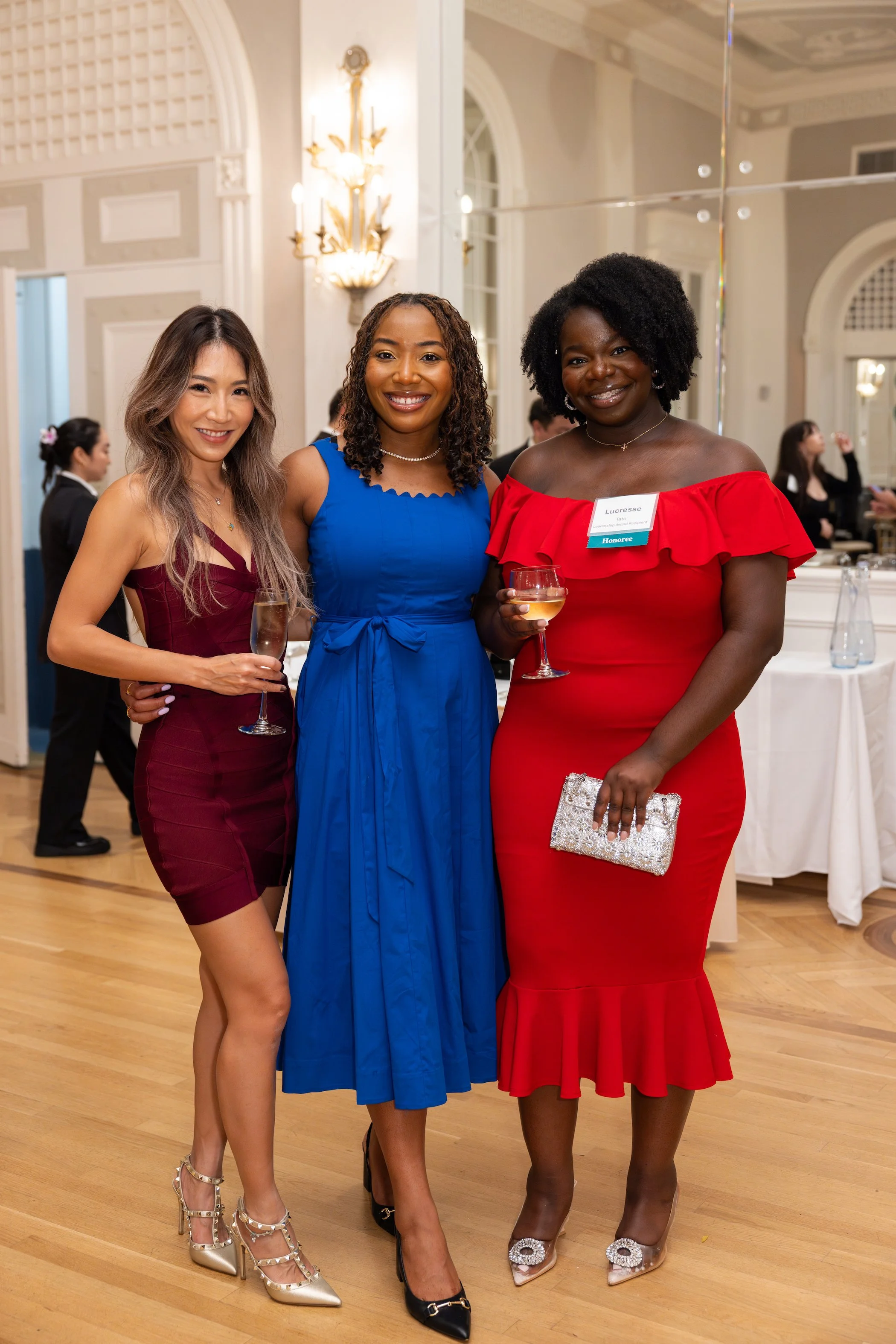 Three women in formal dresses holding glasses of wine at an indoor event.