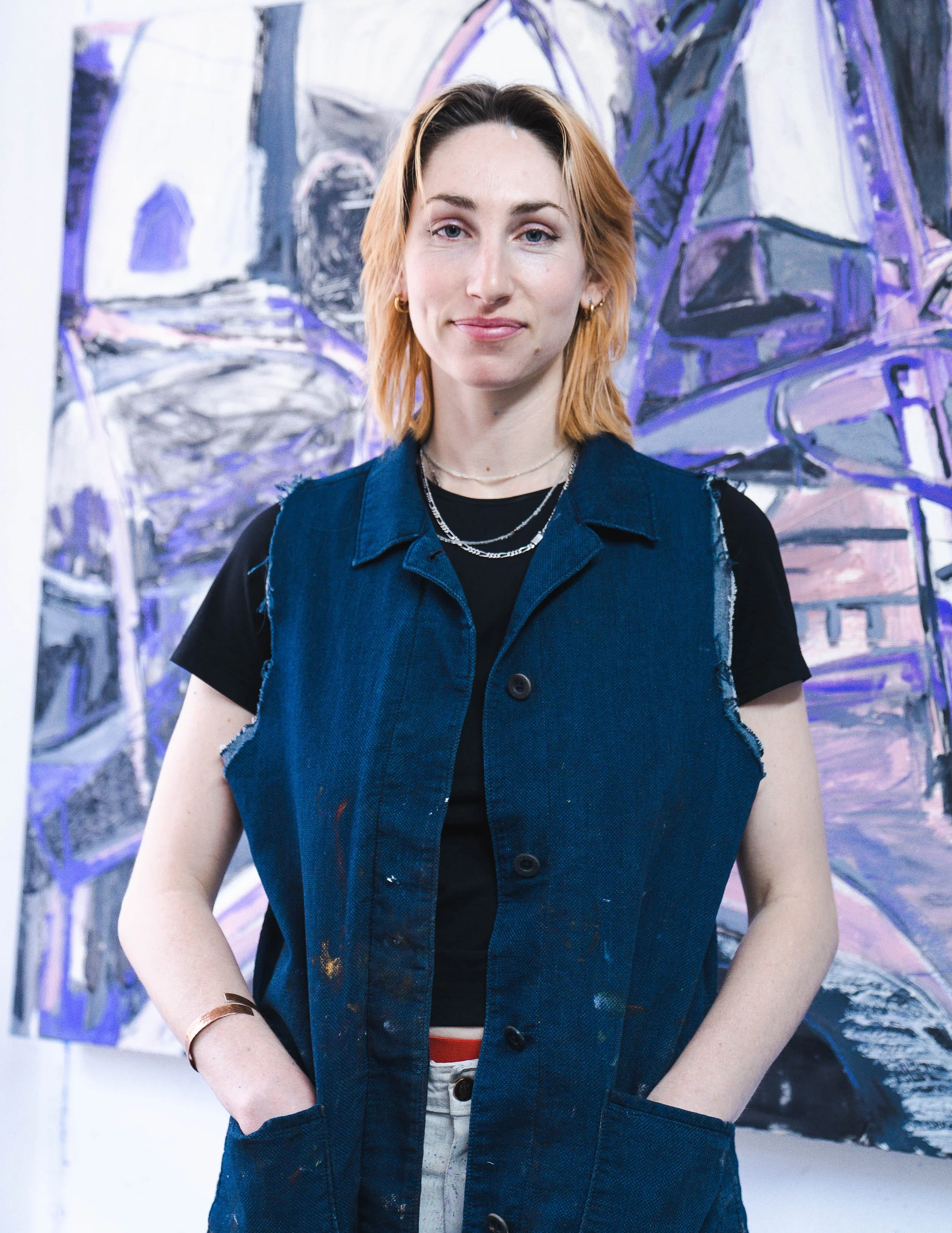 A young woman with shoulder-length reddish-blonde hair, wearing a black t-shirt, layered necklaces, and a sleeveless denim vest, standing in front of an abstract purple and white painting.