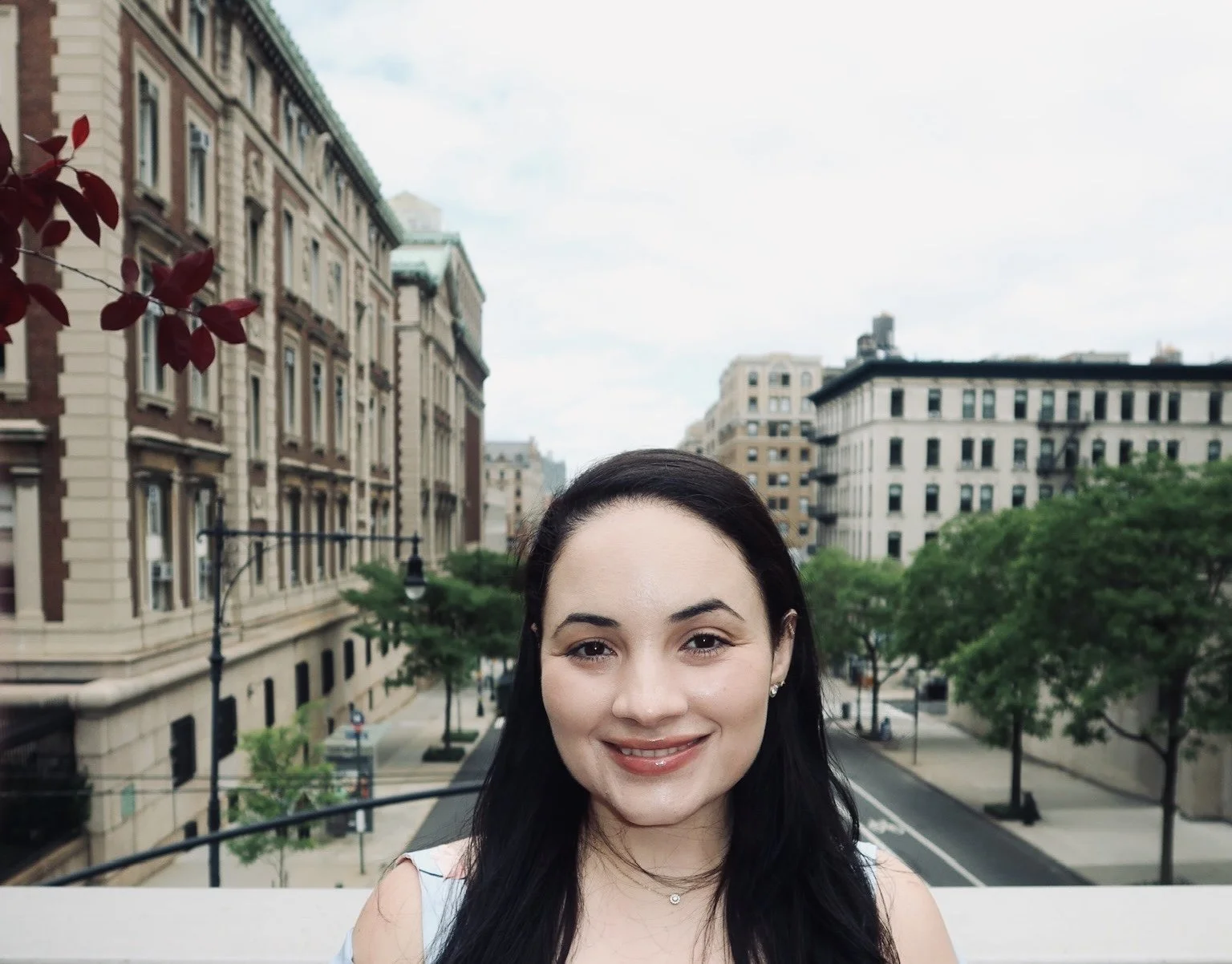 Smiling woman with dark hair standing outdoors on a city street with historic buildings, trees, and overcast sky in the background.