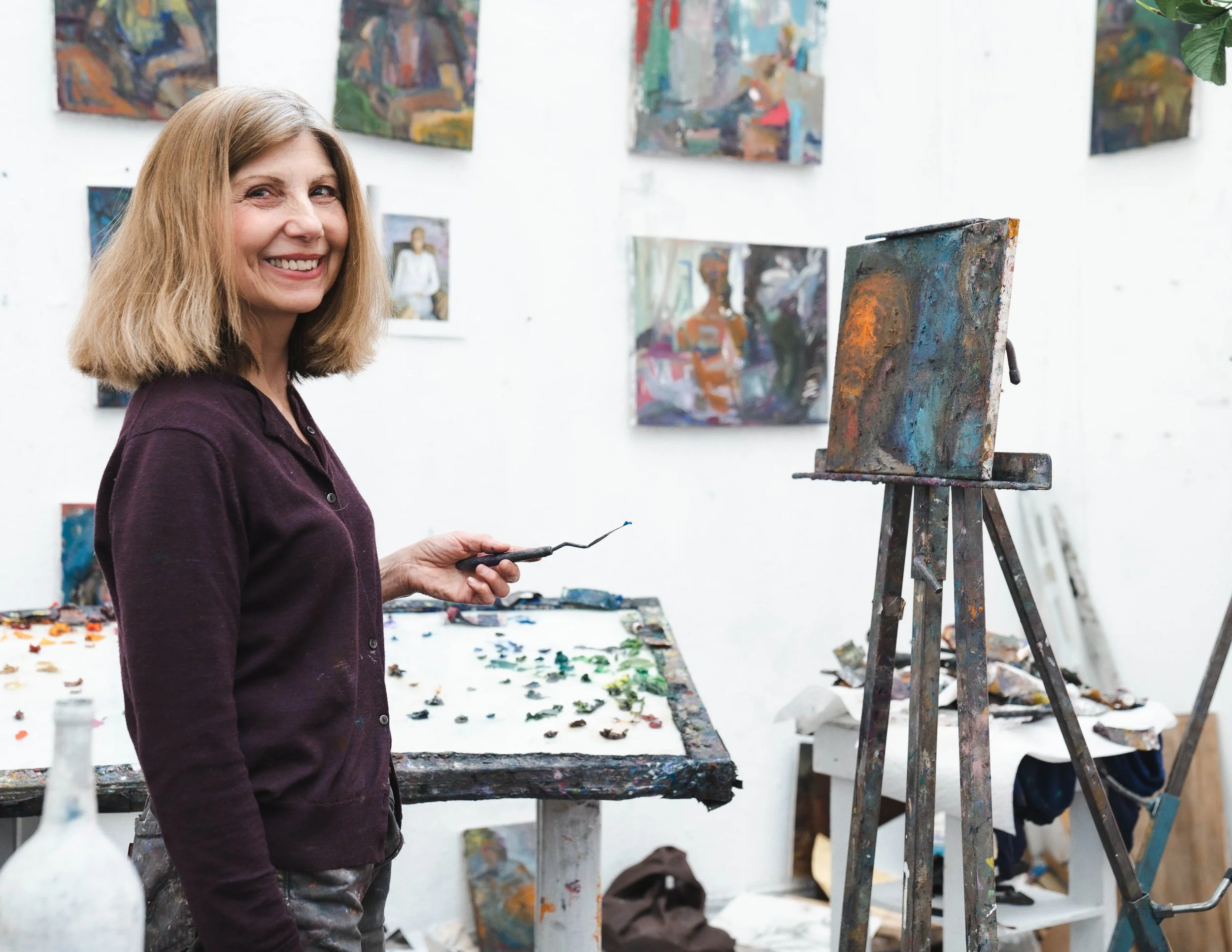 A woman in a purple jacket smiling while holding a paint palette in her hand in an art studio. Behind her are paintings on the wall and an easel with a colorful abstract painting. There are art supplies and painted palettes on the table.