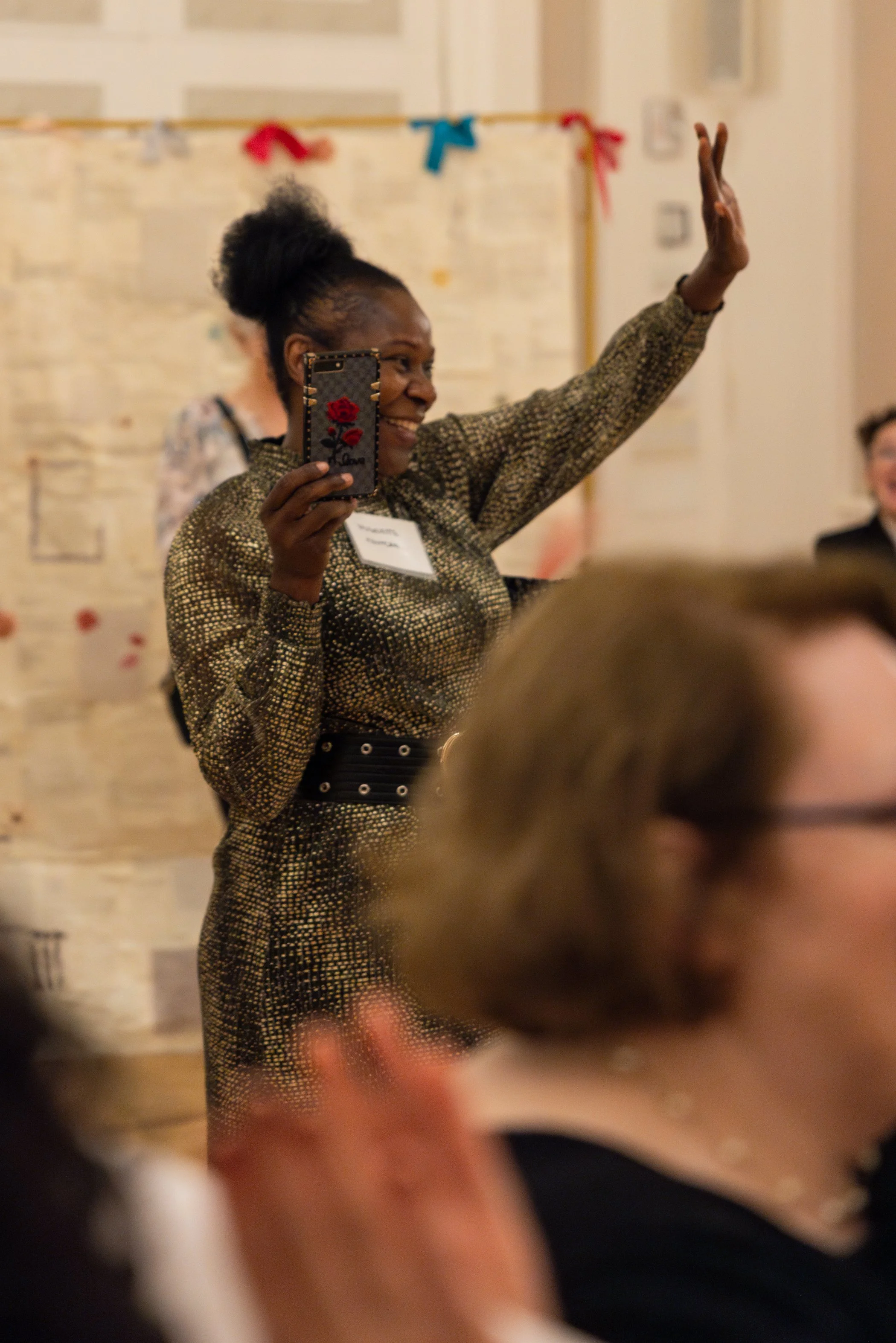A woman with her hair in a bun, wearing a patterned dress with a belt, is smiling and waving with one hand while holding a phone in the other at an indoor event. Other attendees are present in the background.