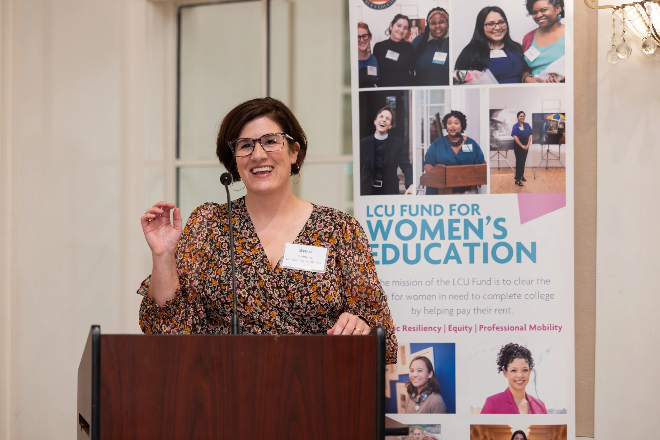 A woman with short brown hair and glasses speaking at a podium, wearing a floral blouse, with a microphone headset. Behind her is a banner that reads 'LCU Fund for Womens Education' and features pictures of diverse women.