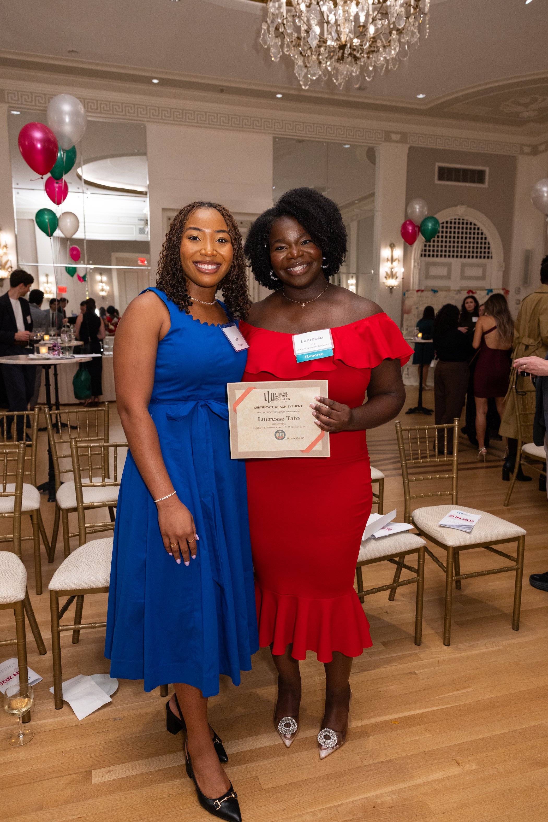 Two women at an awards ceremony, one in a blue dress and the other in a red dress, holding a certificate. Elegant event setting with chairs, balloons, and a chandelier.