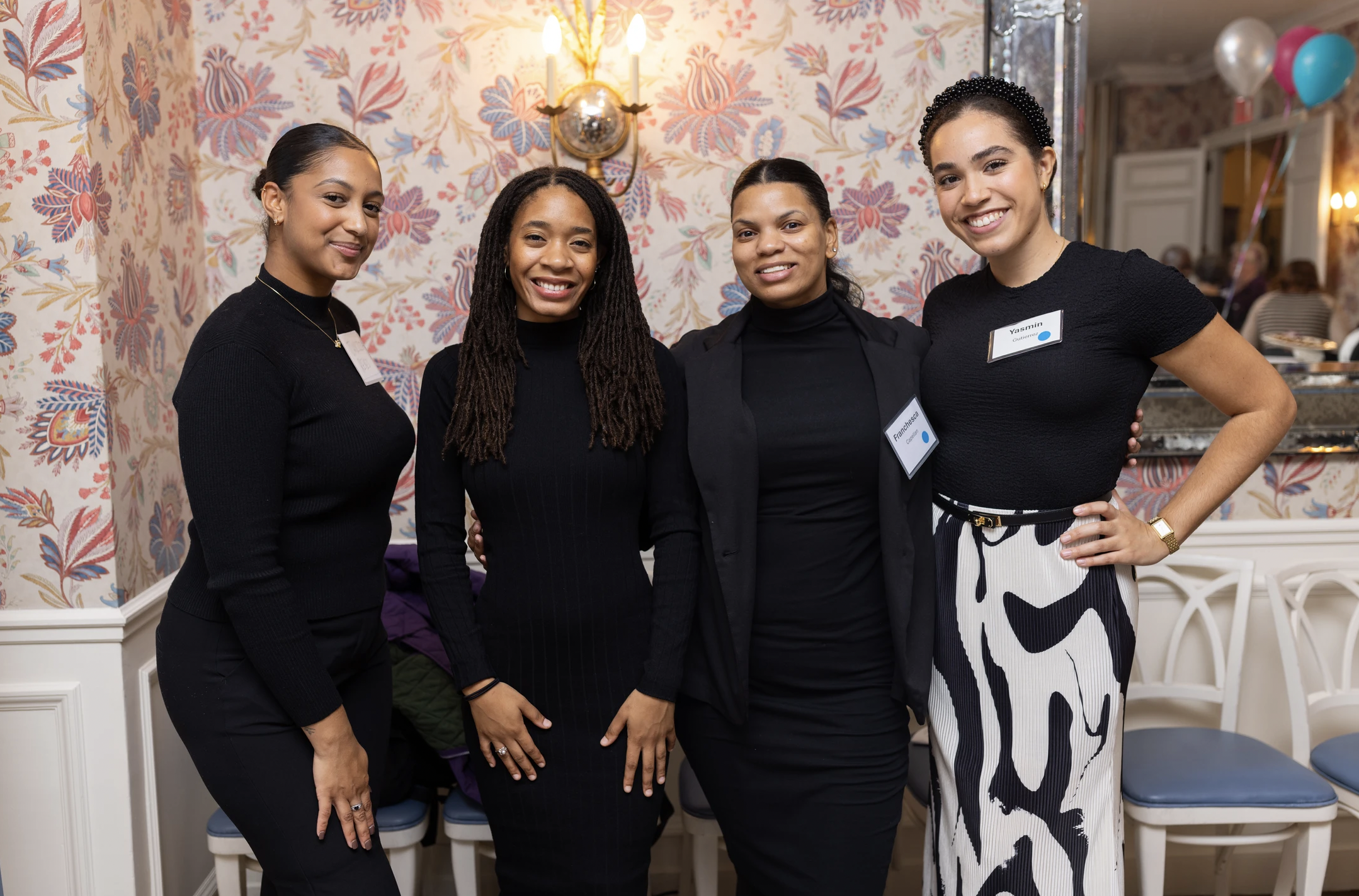 Four women standing together inside a decorated room, smiling at the camera. They are wearing black tops, and one woman has a black and white patterned skirt. The background features floral wallpaper, a wall sconce, balloons, and other people in the 