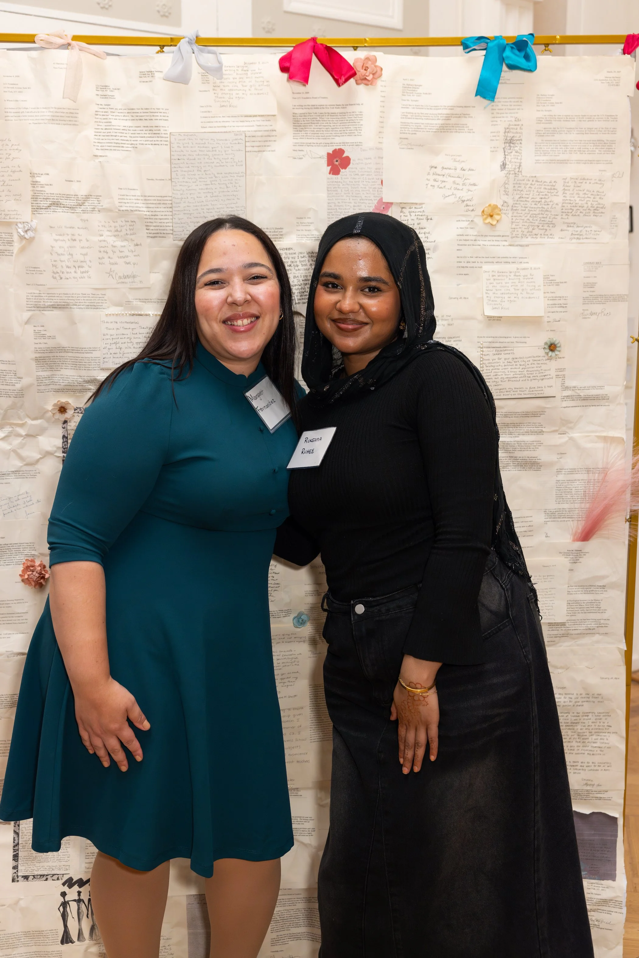 Two women smiling and standing close together in front of a wall covered with papers and colorful bows.