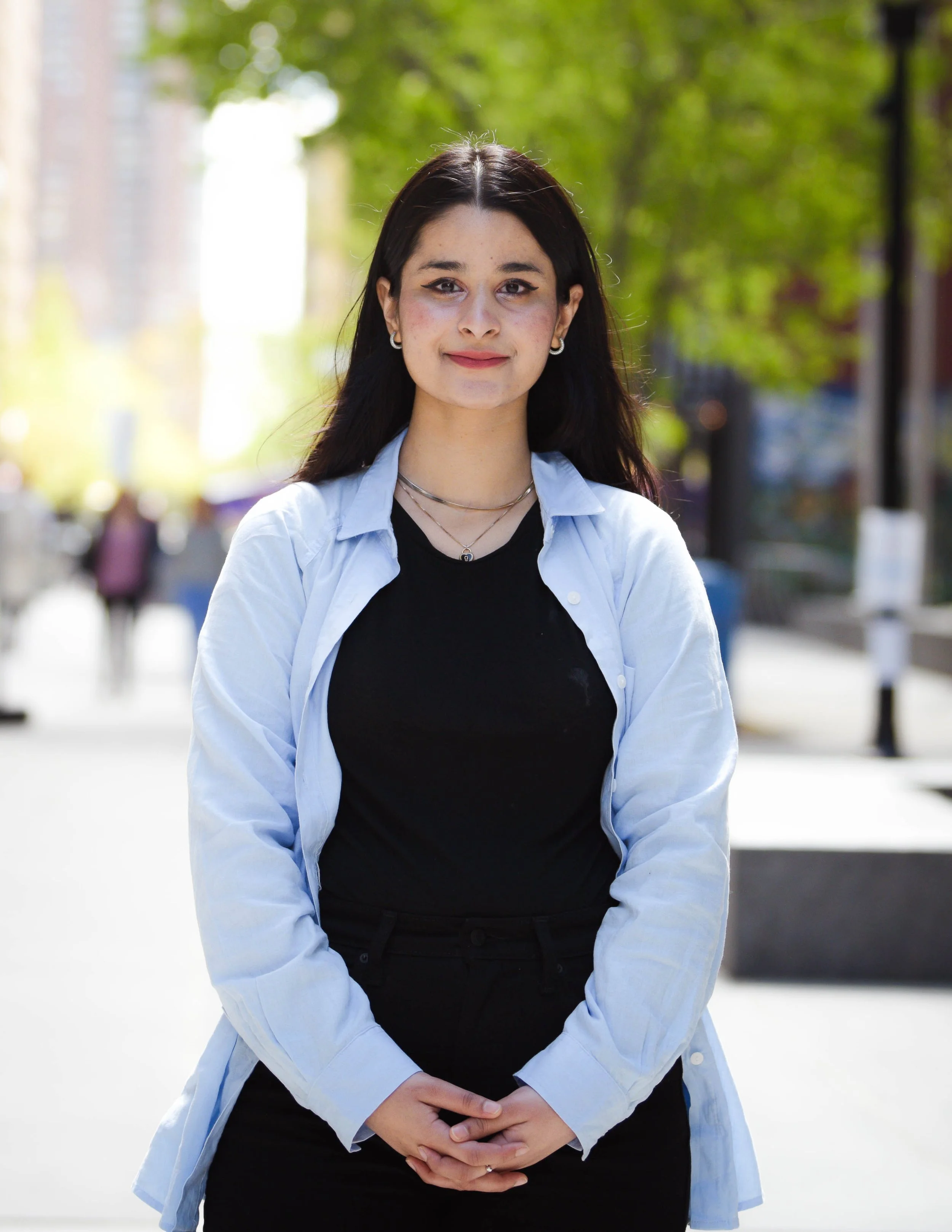 A young woman with dark hair, wearing a black top and a light blue shirt, standing outdoors on a city street with blurred pedestrians and green trees in the background.