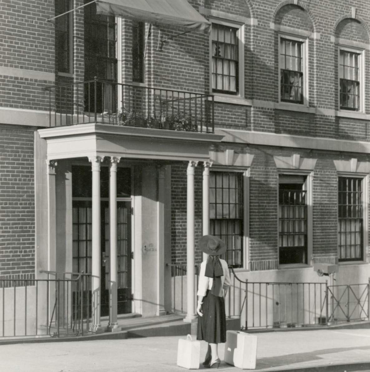 A woman standing outside a brick apartment building with a hat and suitcase, holding a bag, in black and white photo.