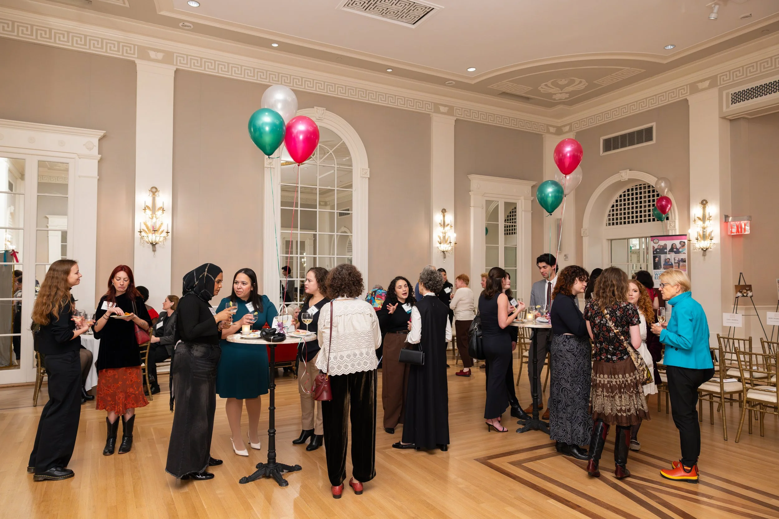 People attending a social event in a spacious, elegant room with balloons, high ceilings, and large mirrors, engaging in conversations and holding drinks.