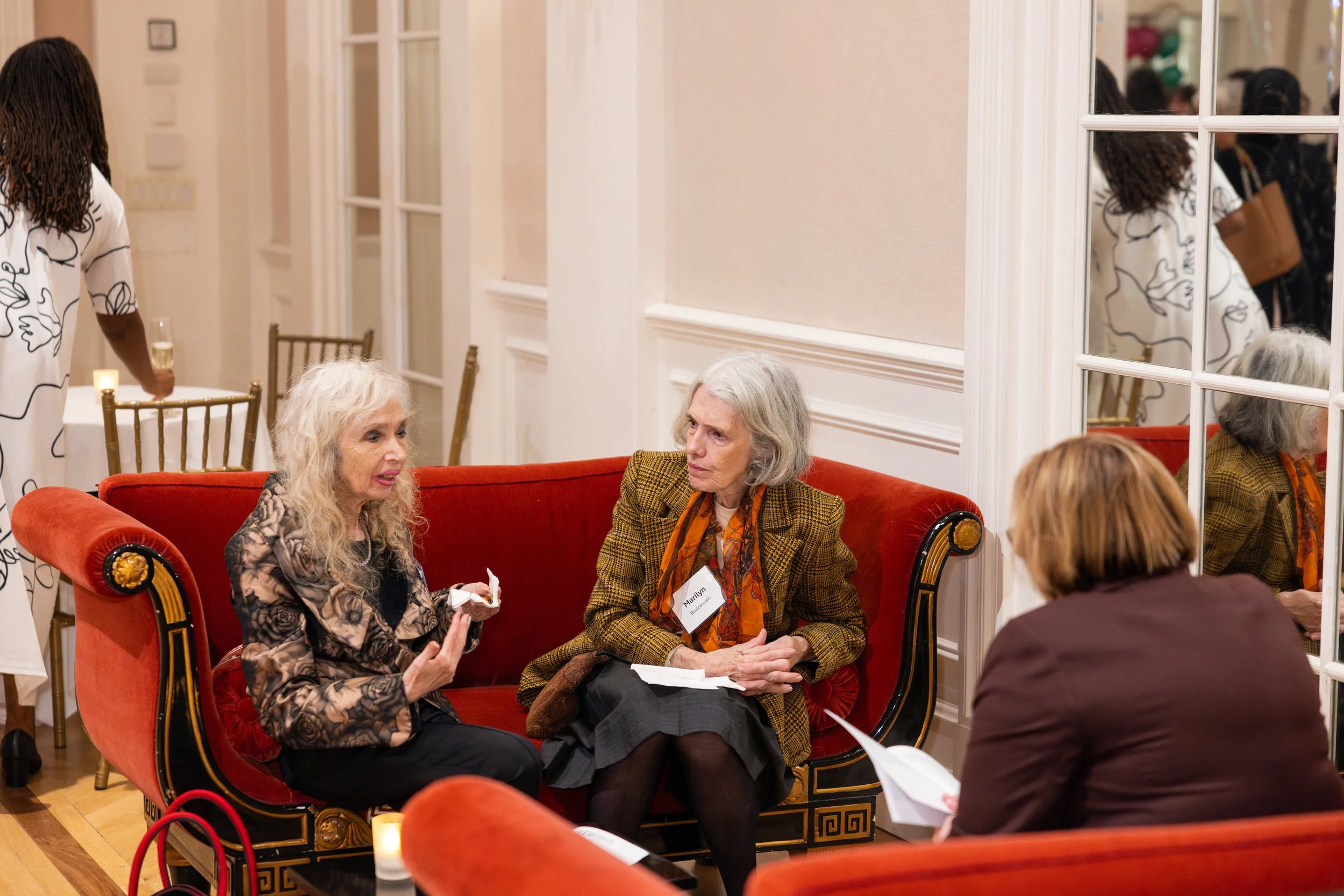 Three women are sitting on a red sofa in a formal gathering or event, engaging in conversation. One woman has long curly white hair and is wearing a patterned jacket, another has short gray hair and is dressed in a tartan blazer with an orange scarf,