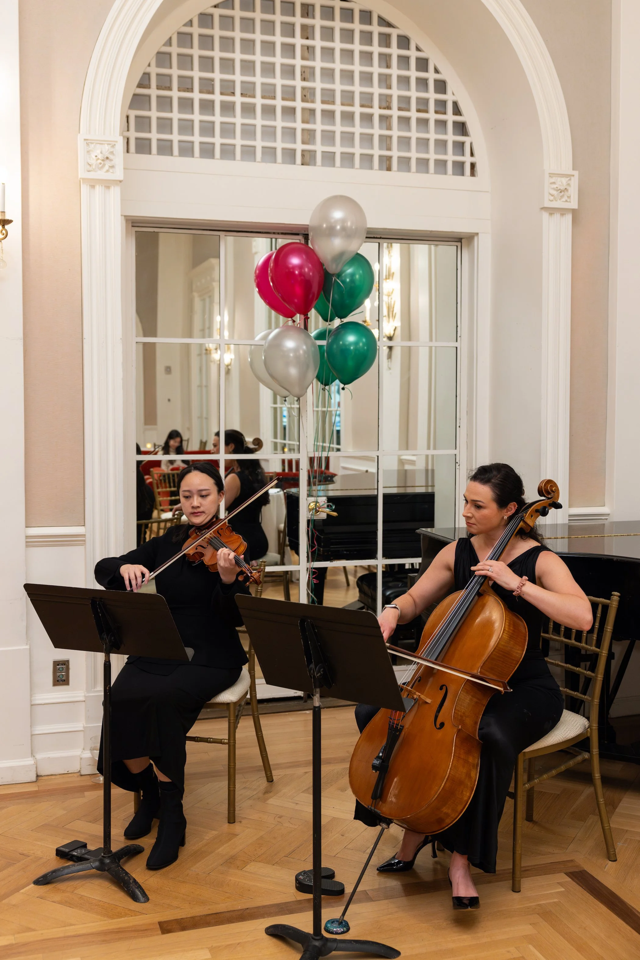 Two women playing string instruments, a violin and a double bass, during a musical performance in a formal setting, with balloons in the background.