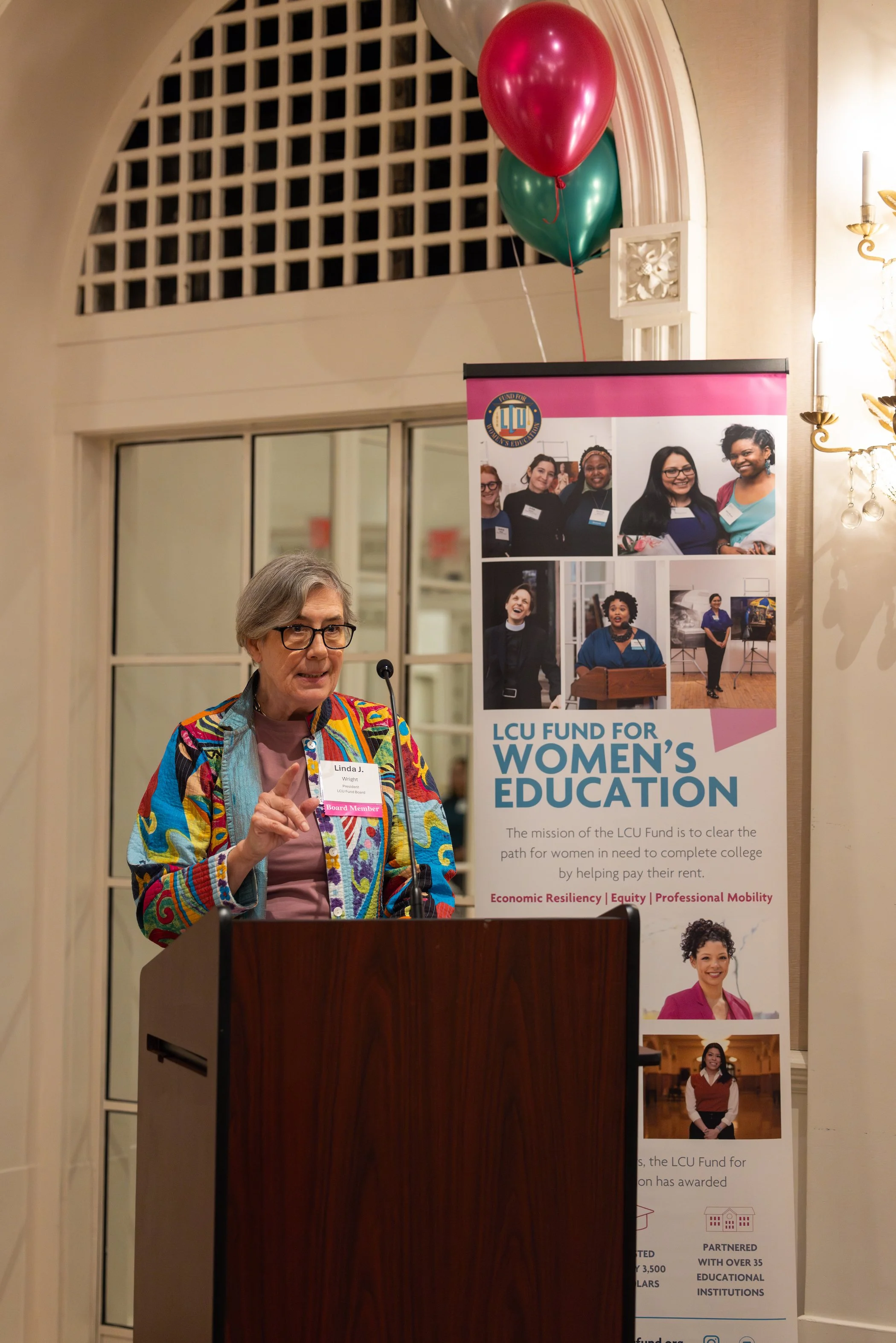 An elderly woman with glasses and gray hair, wearing a colorful jacket, speaks at a podium during a Women's Education fundraiser event for the LCU Fund, which is supporting women in completing college. Behind her is a poster with photos of women and 
