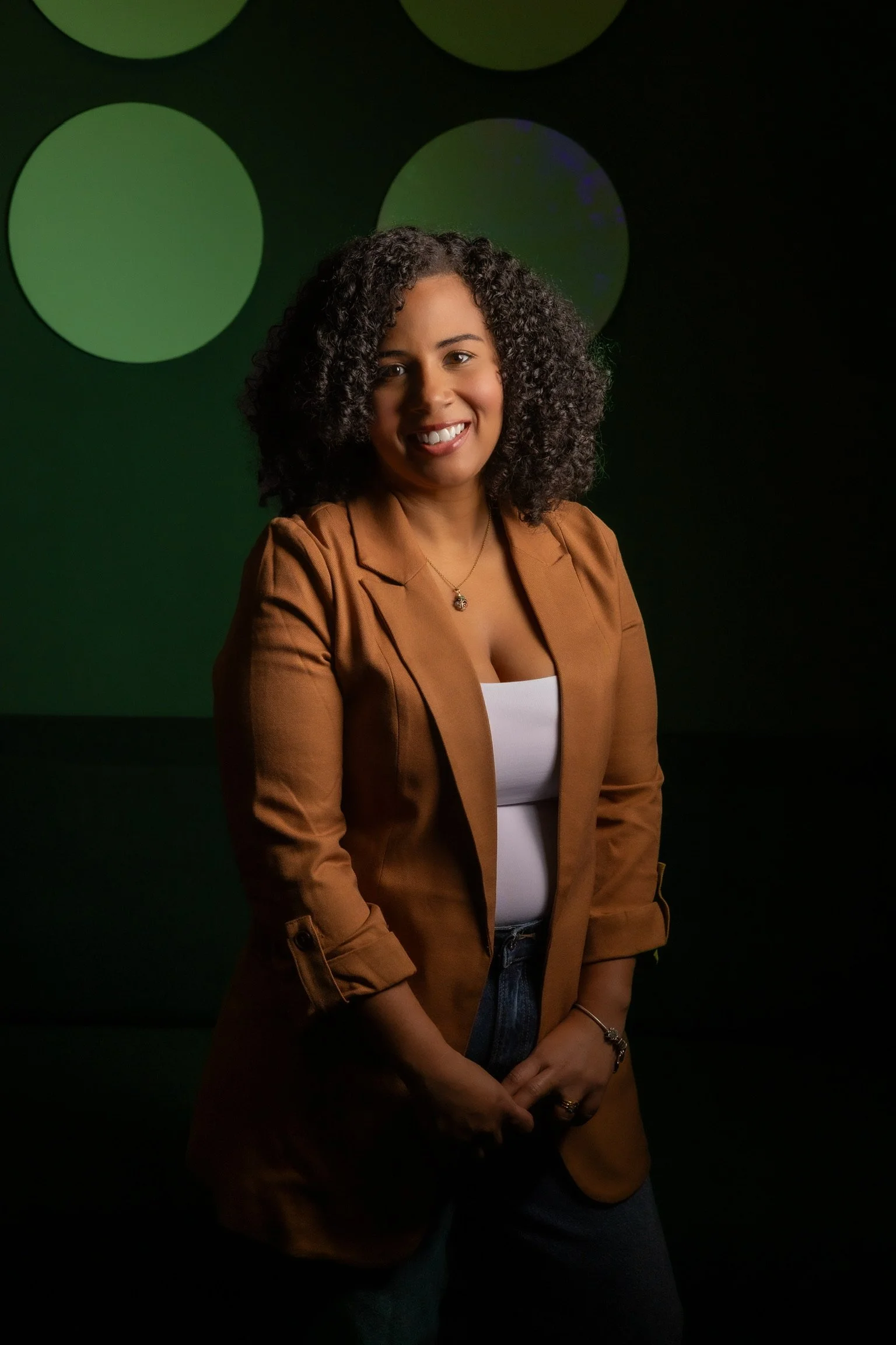 A woman with curly hair, wearing a brown blazer over a white top, standing against a dark background with large green circles, smiling at the camera.