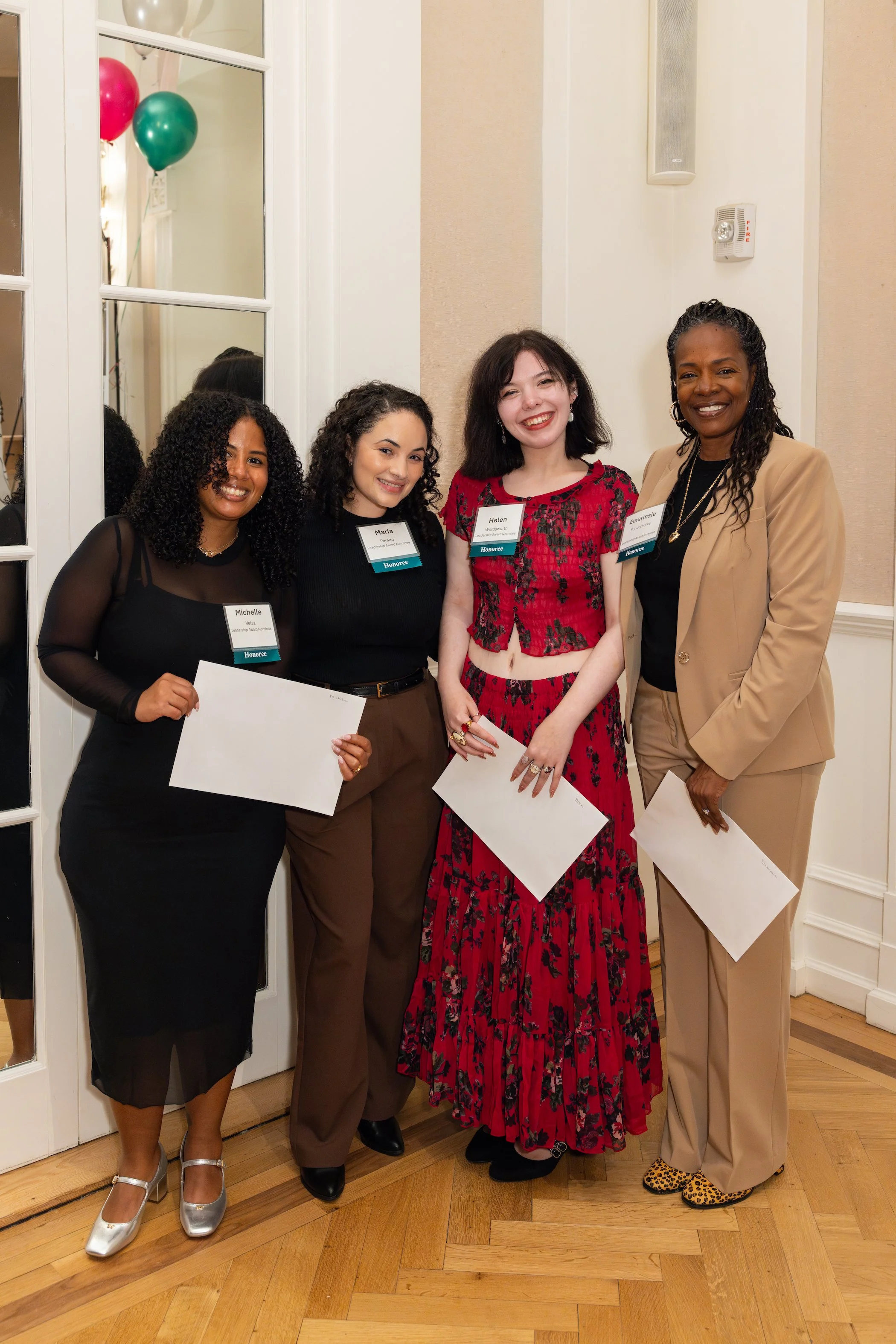 Four women standing side by side, smiling at the camera, holding certificates, at an indoor event with balloons reflected in a mirror behind them.