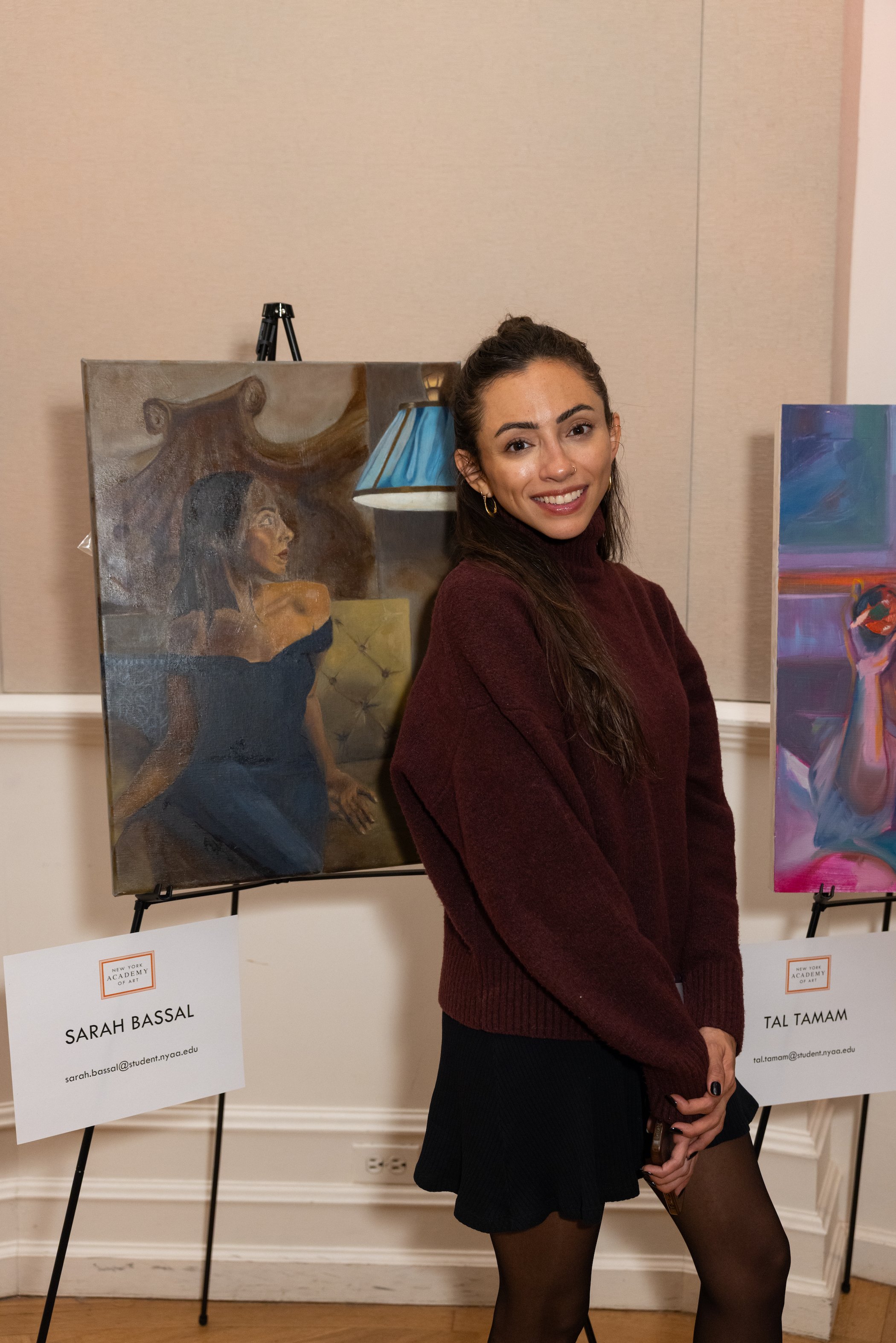 A woman with long brown hair in a bun, wearing a maroon sweater, standing beside two framed paintings on easels, in an art gallery.