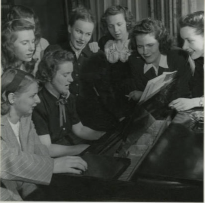 A group of women gathered around a piano, with one woman playing and the others singing.