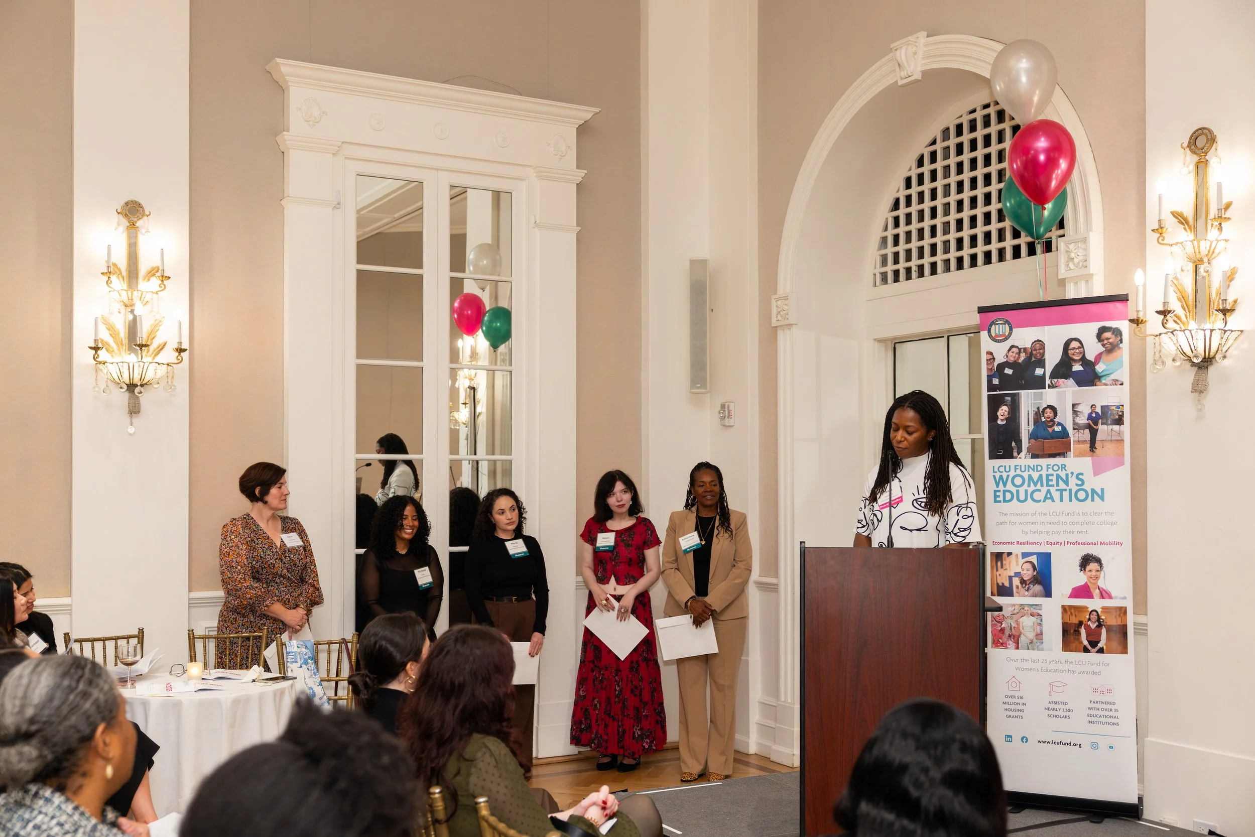 A woman speaking at a podium during a women's education event, with a panel of diverse women standing behind her, in an elegant room with high ceilings, large mirrors, and balloons.