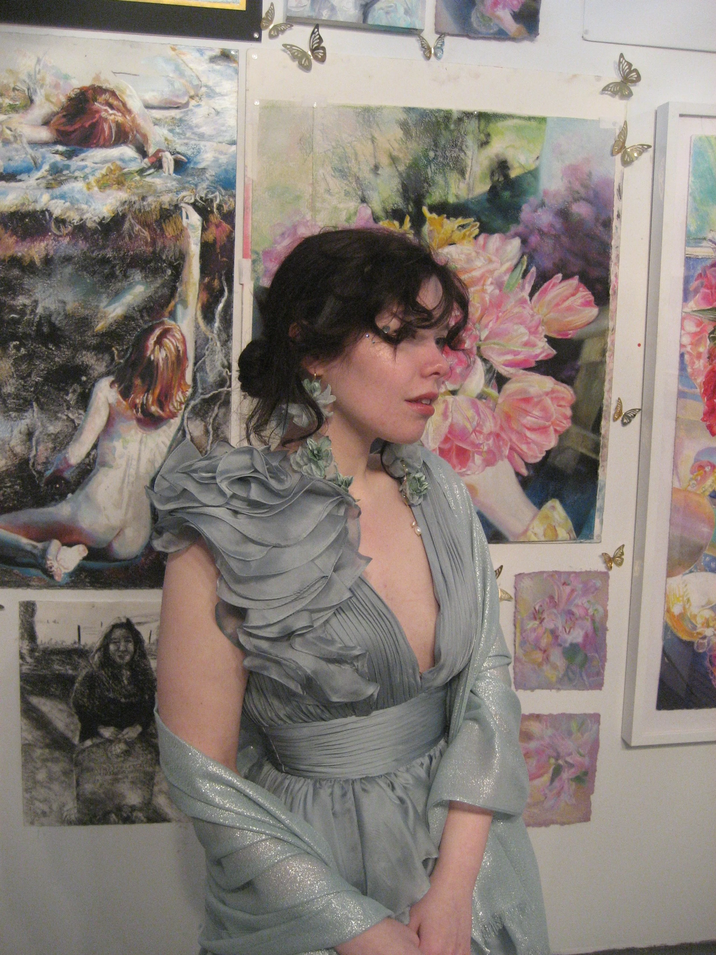 A woman in a silver dress with ruffled shoulder details poses in front of colorful floral and artistic paintings on an art gallery wall.