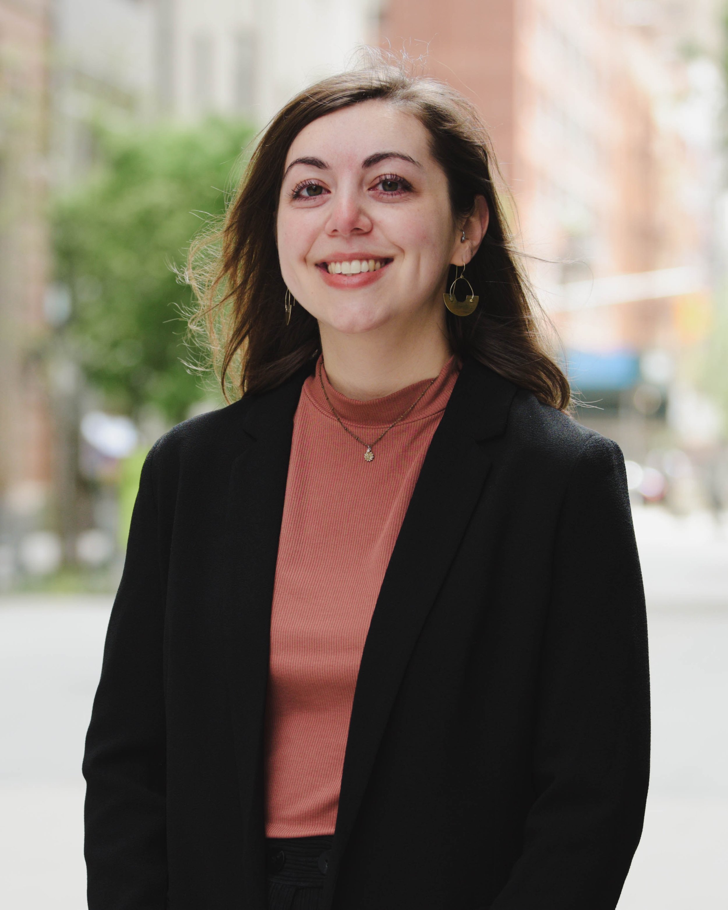 A young woman with brown hair, smiling, wearing a black blazer, blush pink top, and gold jewelry, standing outdoors with blurred trees and buildings in the background.