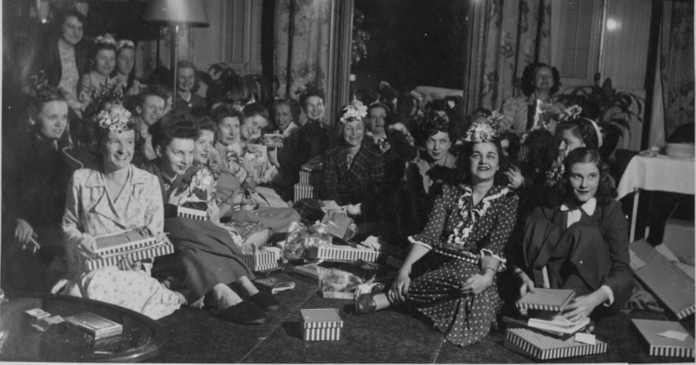 Group of women and girls at a celebration party, some wearing floral hats, opening presents, with boxes and gifts on the table.