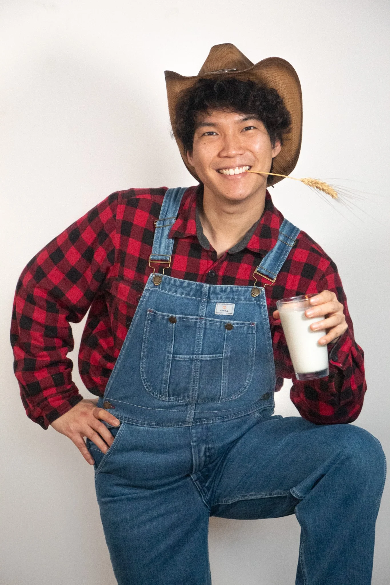 A young man wearing a cowboy hat, red and black plaid shirt, and blue overalls, holding a glass of milk, with a wheat stalk in his mouth, smiling at the camera against a plain white background.