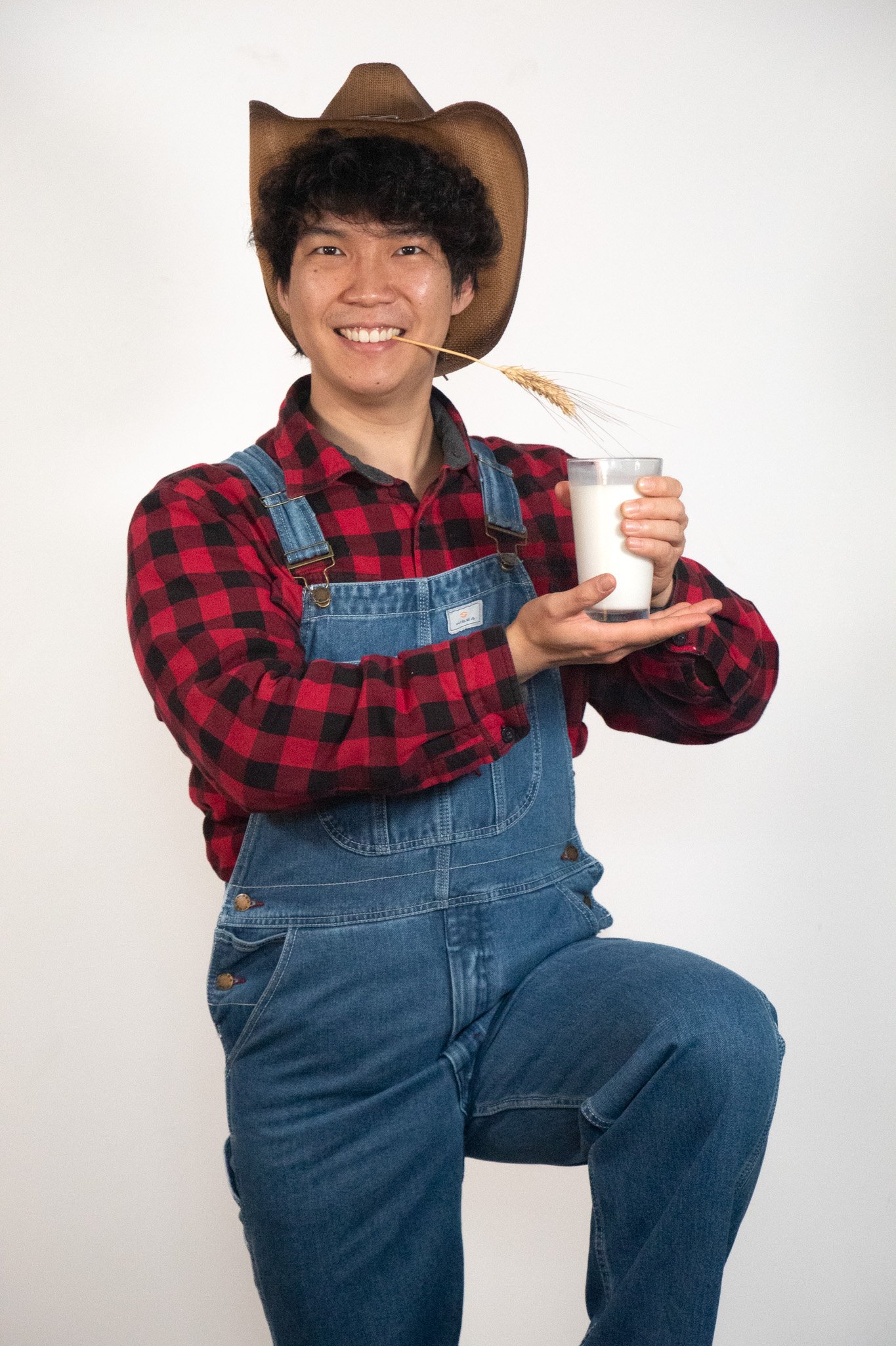 A person wearing a cowboy hat, red and black plaid shirt, and denim overalls, holding a glass of milk and a wheat stalk, smiling at the camera against a plain white background.