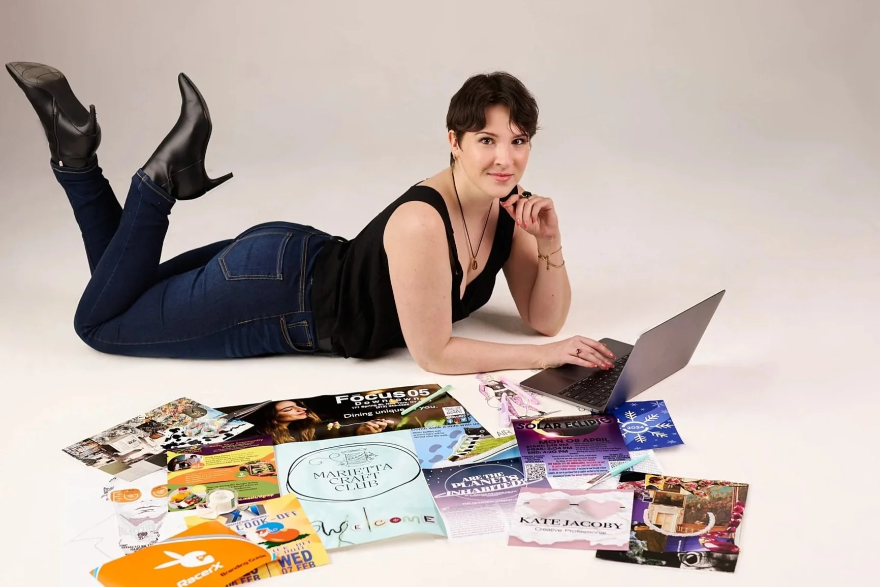 Katie Jacoby is depicted laying on the ground with her laptop with samples of her artwork collaged in the foreground.