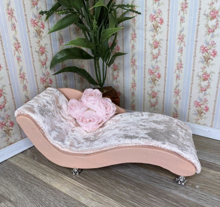 A small pink chaise lounge with velvet fabric, decorated with pink roses, placed on a wooden floor next to a plant with green leaves and a flowered wallpaper background.