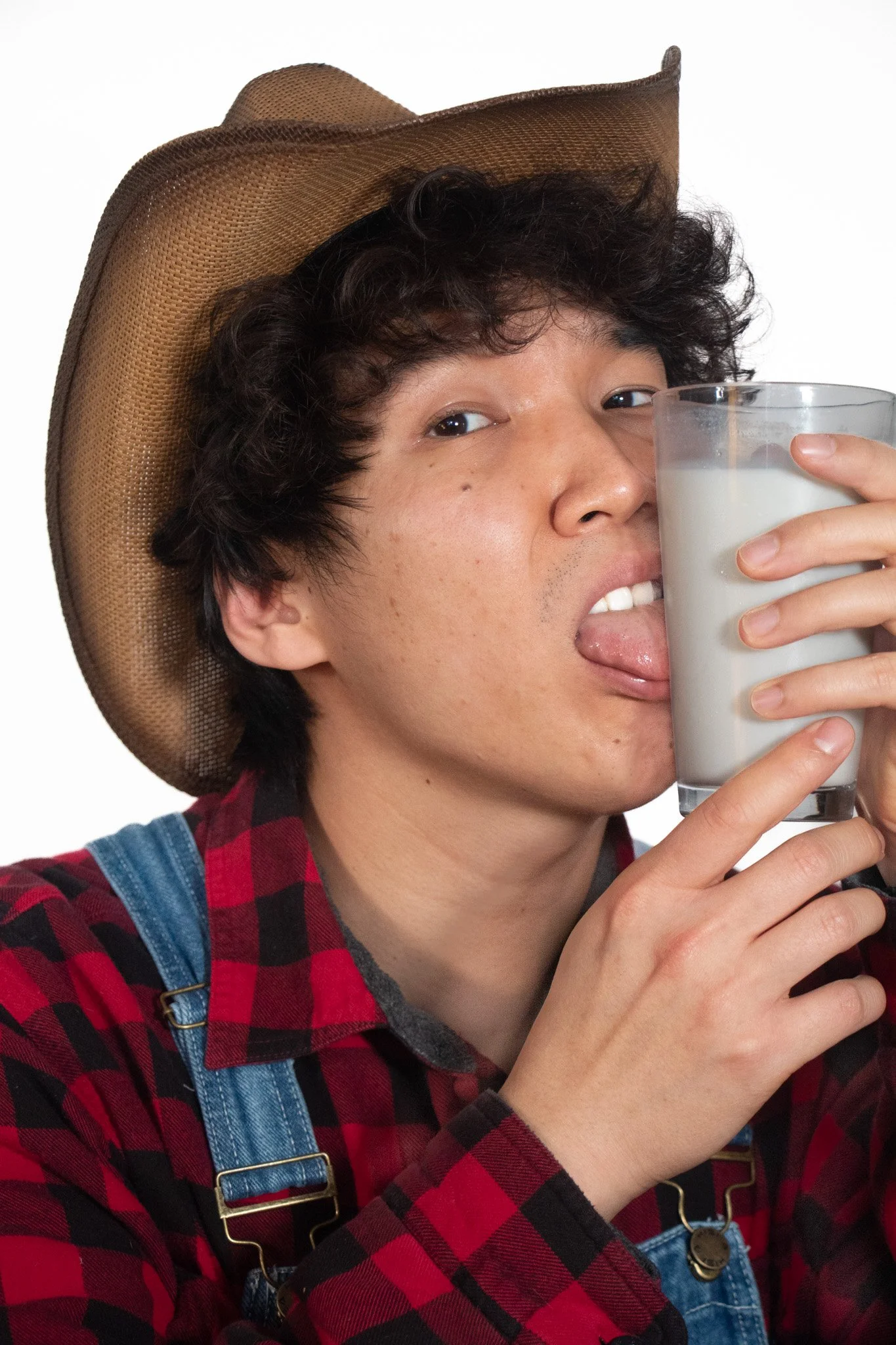 A person wearing a cowboy hat, red and black plaid shirt, and denim overalls, holding a glass of milk near their mouth with their tongue out, against a plain white background.