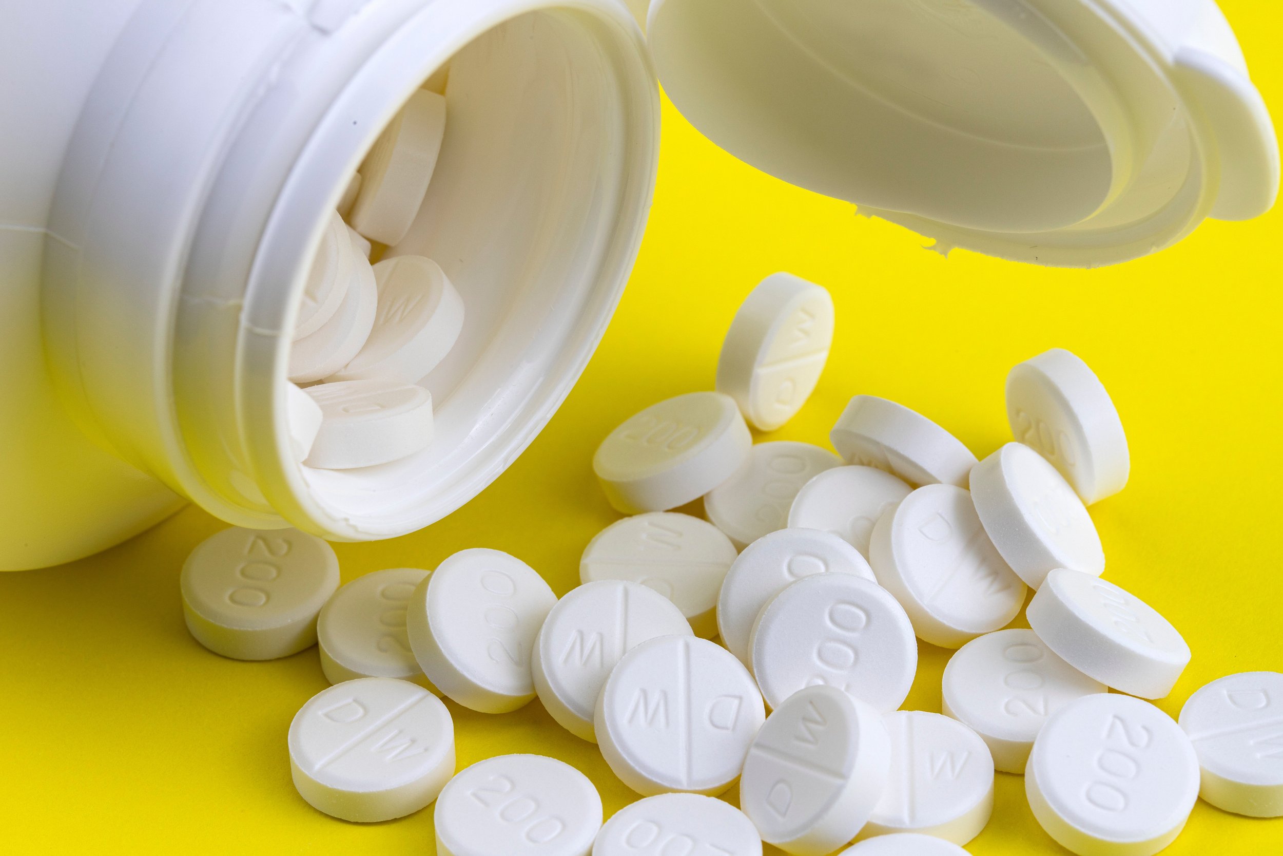 A white pill bottle tipped over with white round pills spilling onto a yellow surface.