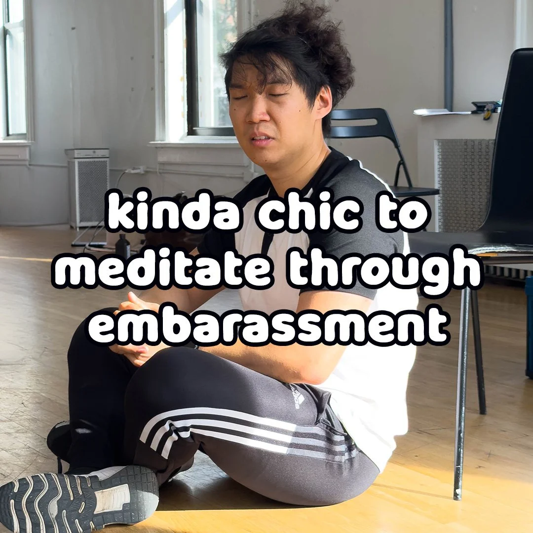Young man with messy hair sitting on the floor with eyes closed, practicing meditation in a bright room with large windows, a chair, and a laptop in the background. Text overlay: 'kinda chic to meditate through embarassment'.
