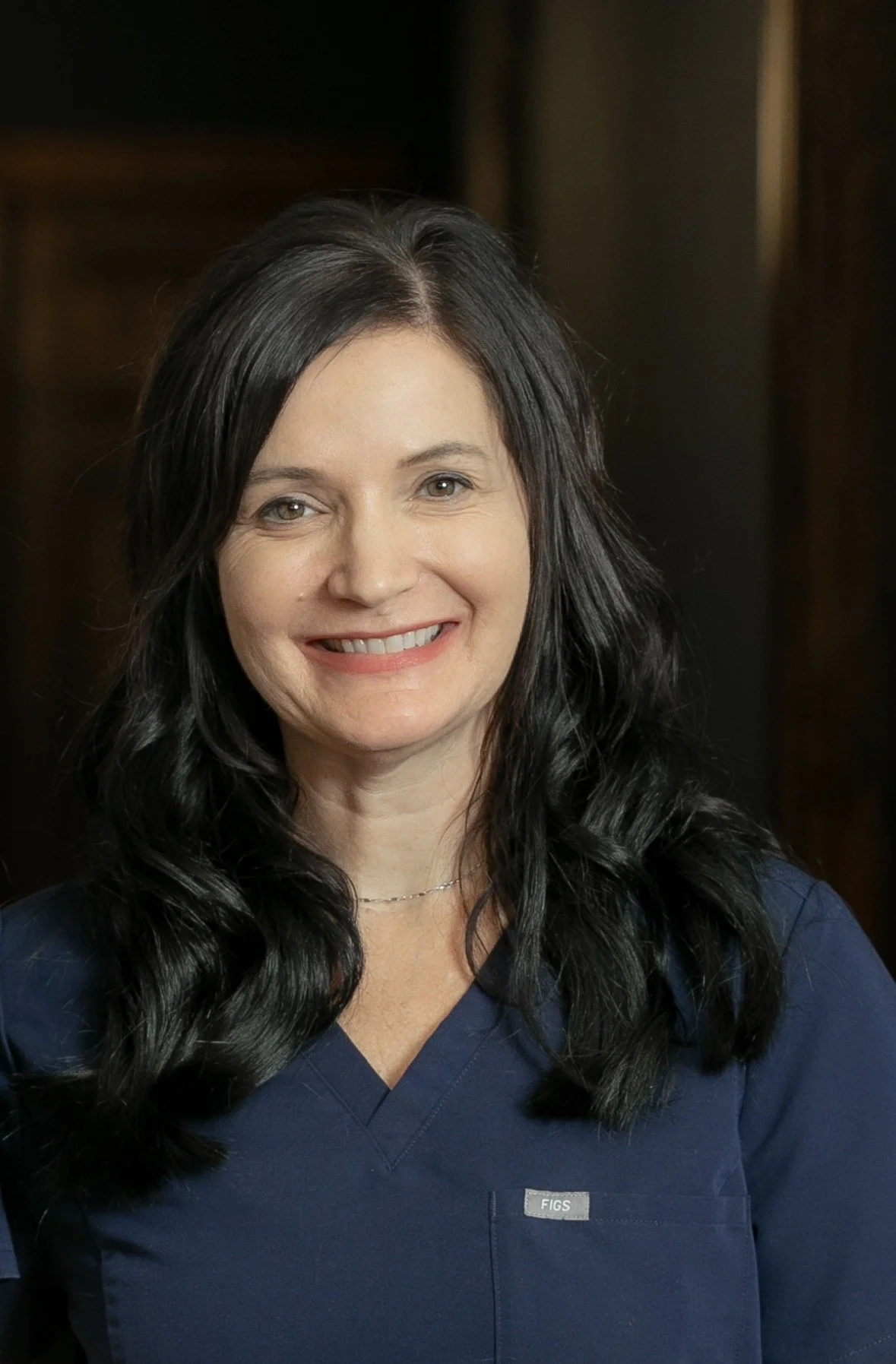 A woman with short dark hair, wearing a black Lake Okeechobee Med Spa t-shirt, smiling indoors in front of a mirror and white brick wall.