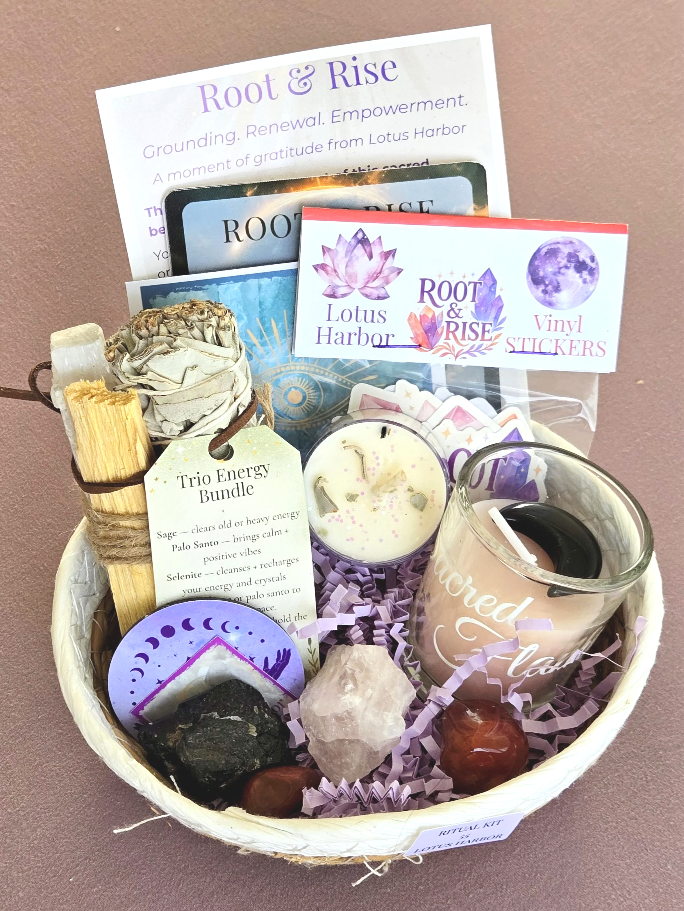 A woven basket containing crystals, gemstones, candles, and wellness items, with decorative cards and flyers about 'Root & Rise' and 'Lotus Harbor' inside and around the basket.