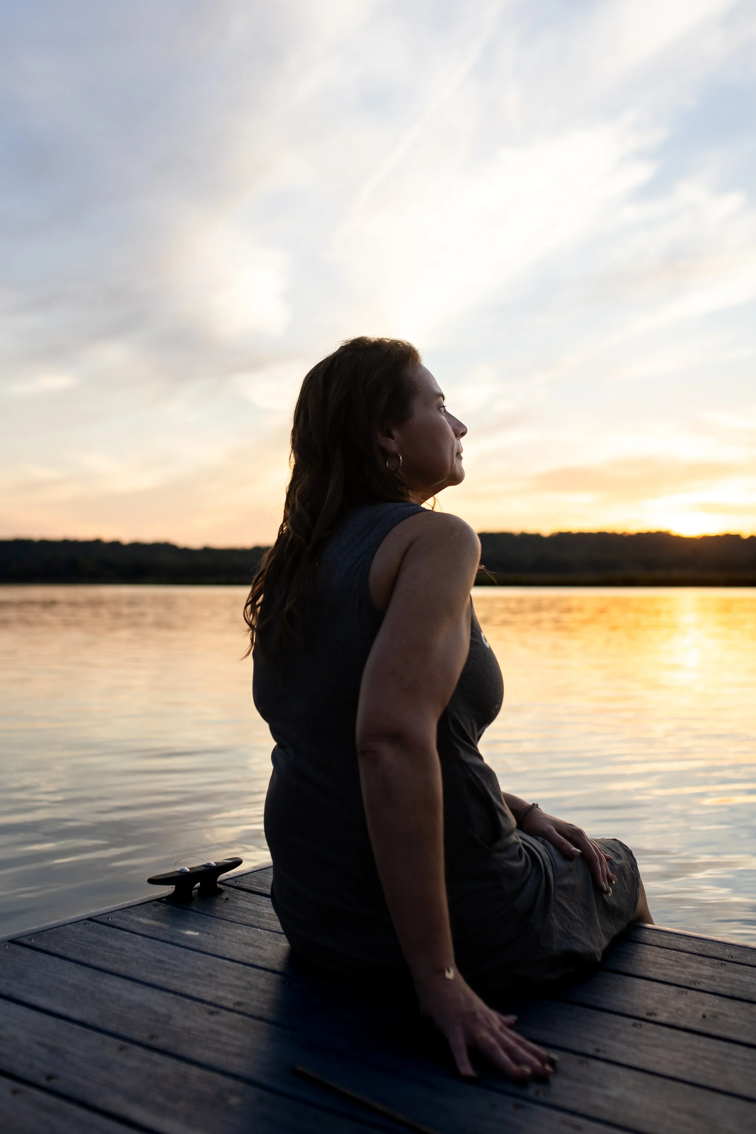 A woman sitting cross-legged on a wooden dock by a body of water, looking at the sunset with clouds in the sky.