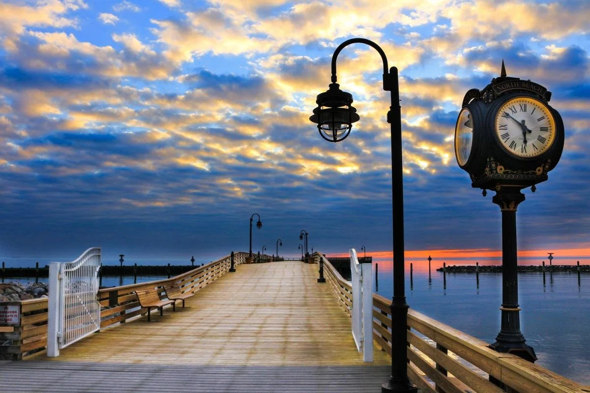 A wooden pier extending into the water at sunset, with benches on the side, street lamps, a clock with Roman numerals, and a cloudy sky with orange hues near the horizon.
