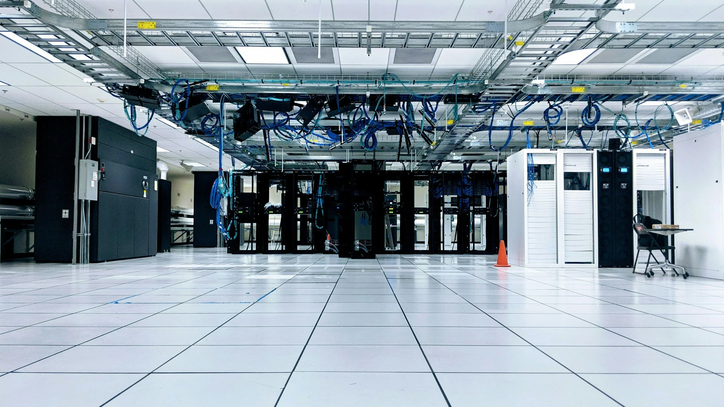 Data center server room with black server racks, network cables hanging from ceiling, large white electrical cabinets, and a person working at a desk.