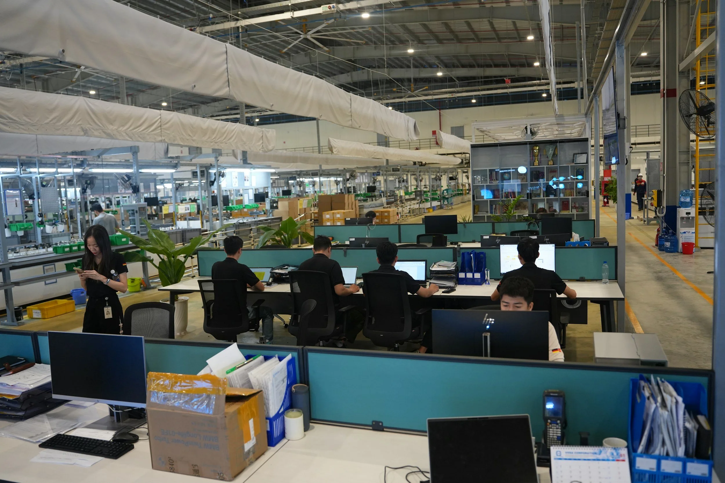 Inside a large industrial warehouse with workers and multiple computer workstations, shelves with awards and books, and a conveyor belt system overhead.