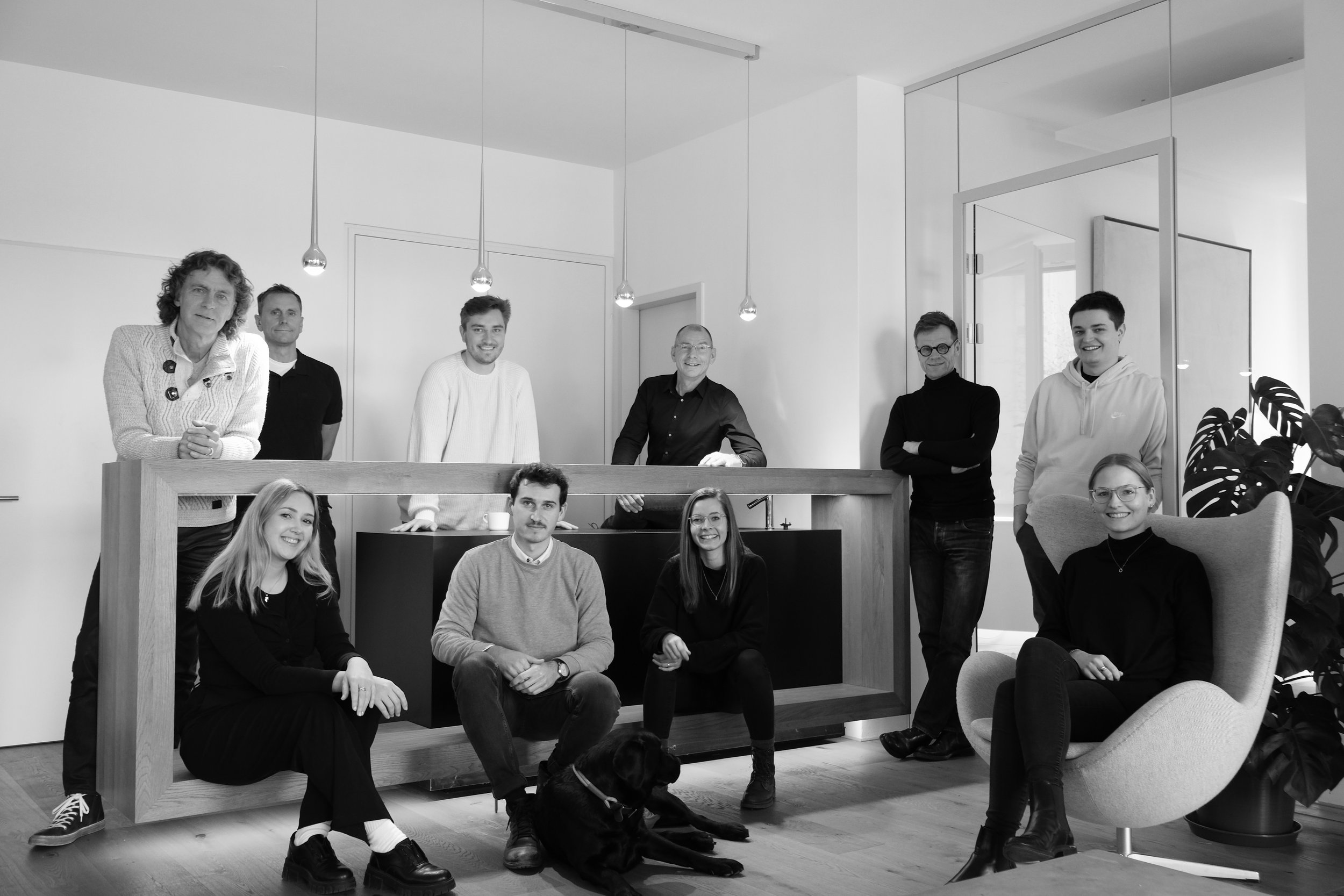 Gruppe von neun Menschen und einem Hund in einem modernen Raum, einige sitzen, andere stehen, alle lachen oder lächeln, im Hintergrund ein Raumtrennwand und Pflanzen.