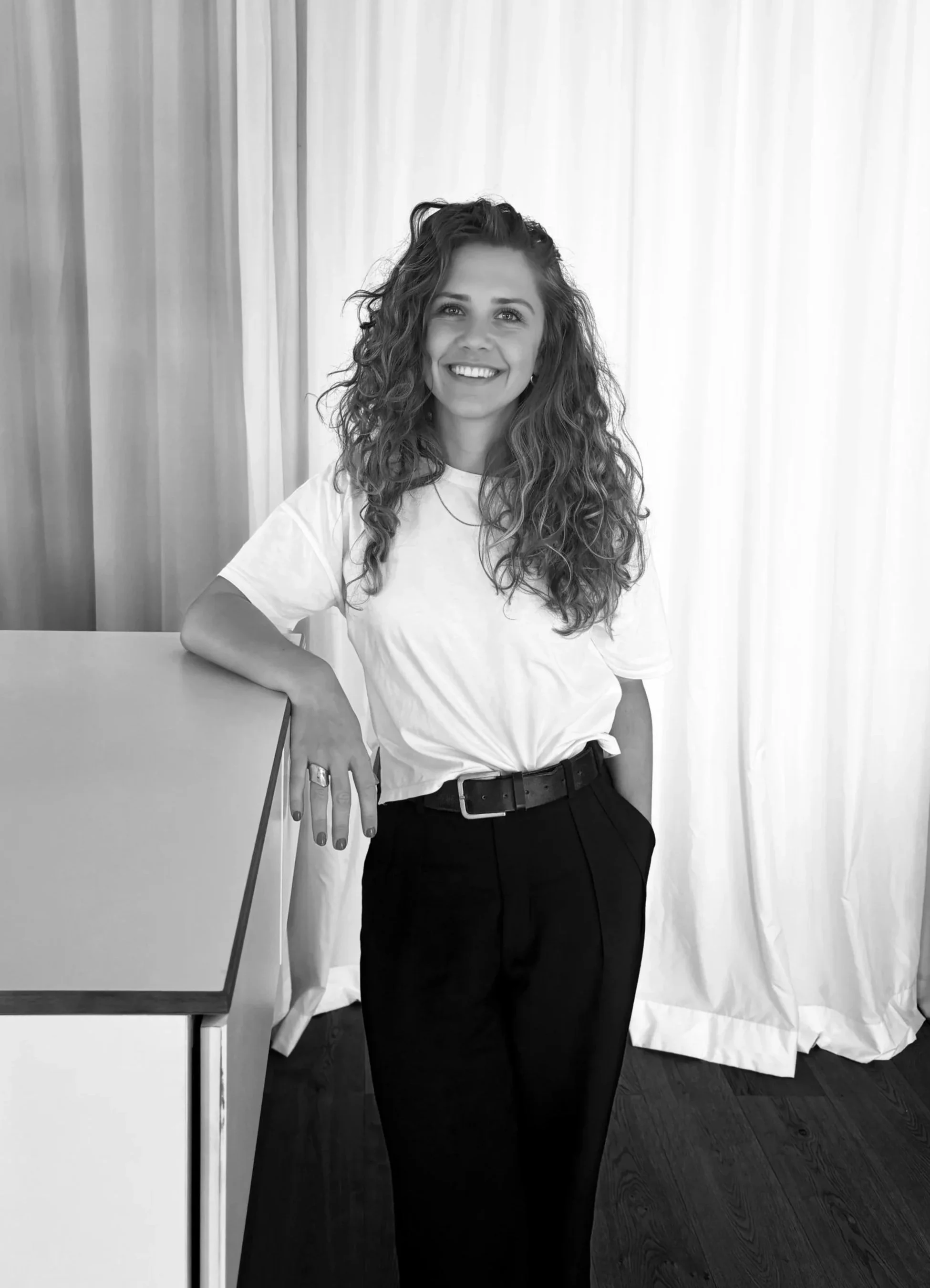 Junge Frau mit lockigem Haar, weißes T-Shirt und dunklen Hosen posiert lächelnd vor einer weißen Stoffhintergrundu.