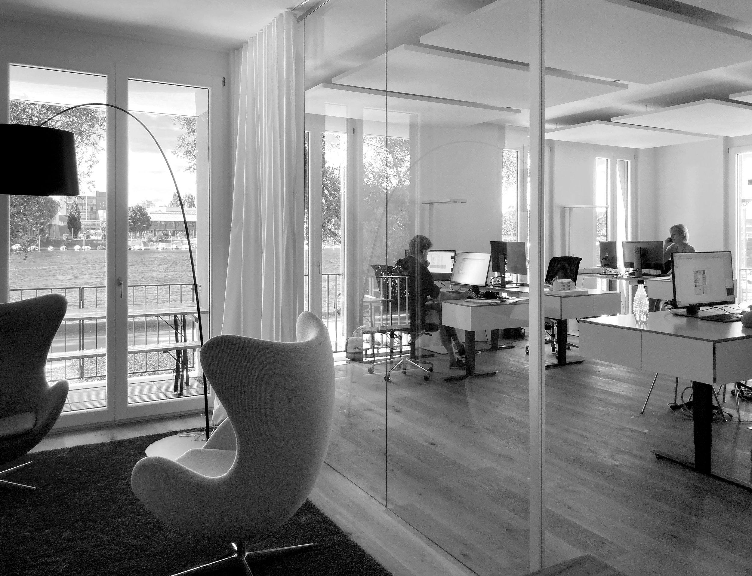 Ein modernes Büro mit großen Fenstern, mehreren Arbeitsplätzen mit Computern, einer modernen Steh-Sitz-Ergonomie-Stuhl und einem Blick auf eine Wasserstraße im Hintergrund.