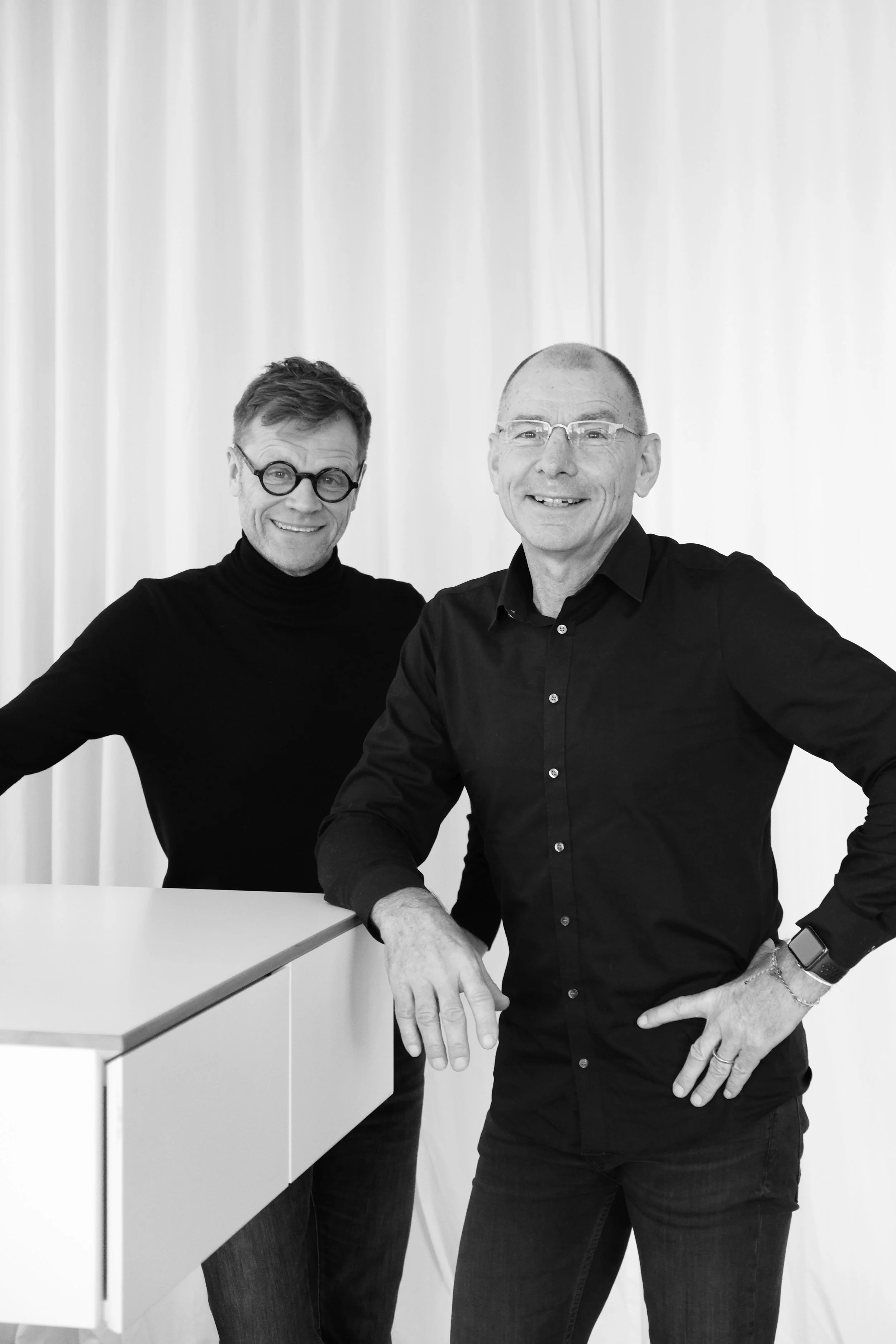 Zwei Männer in schwarzen Hemden und Brillen posieren vor einem weißen Hintergrund.
