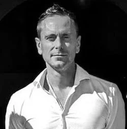 Schwarz-weiß-Foto eines Mannes, der einen weißen Hemd trägt, mit kurzen Haaren, vor dunklem Hintergrund.