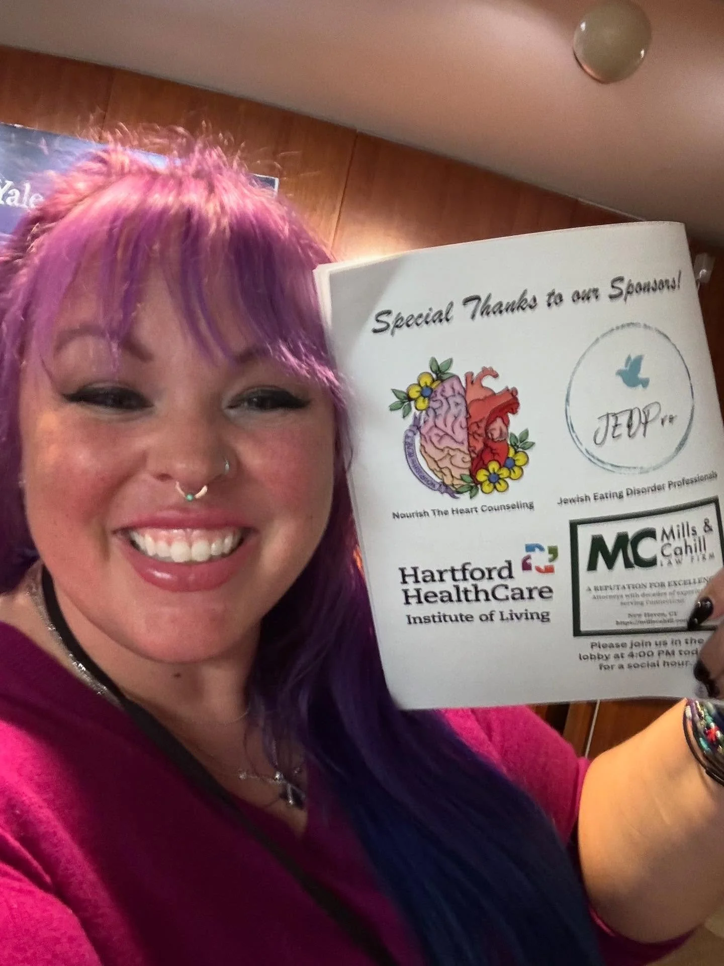 A woman with purple and pink hair, smiling, holding a flyer for a health care event, with logos for Hartford HealthCare and other sponsors.