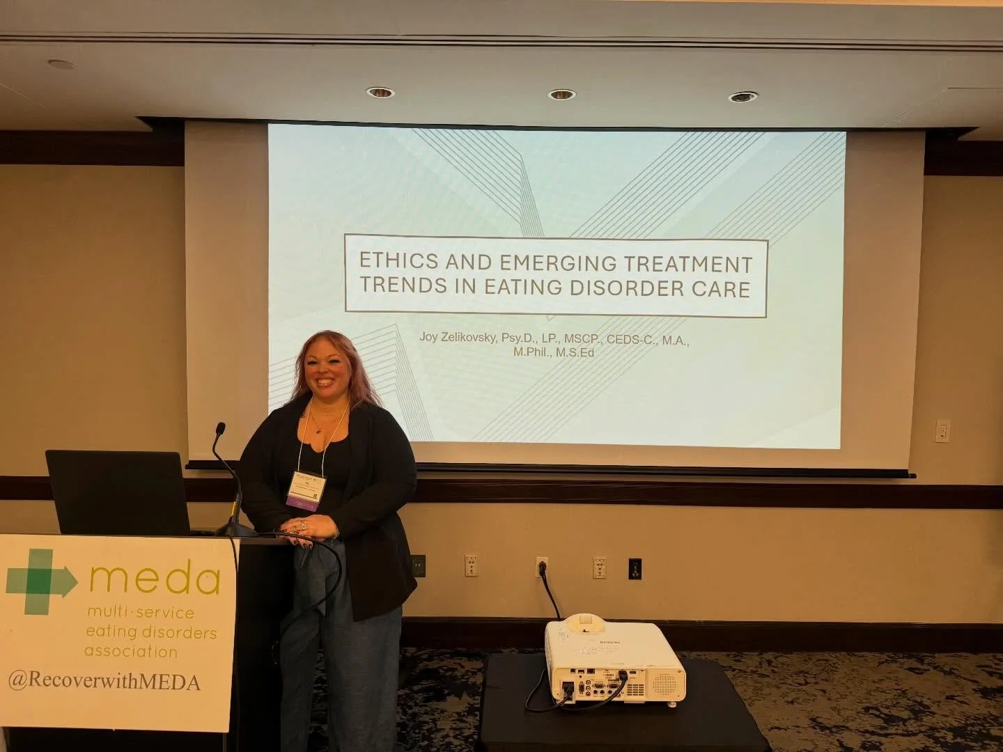 A woman standing at a podium with a laptop, smiling, during a presentation on ethics and emerging treatment trends in eating disorder care, with a screen behind her displaying the presentation title and her credentials.