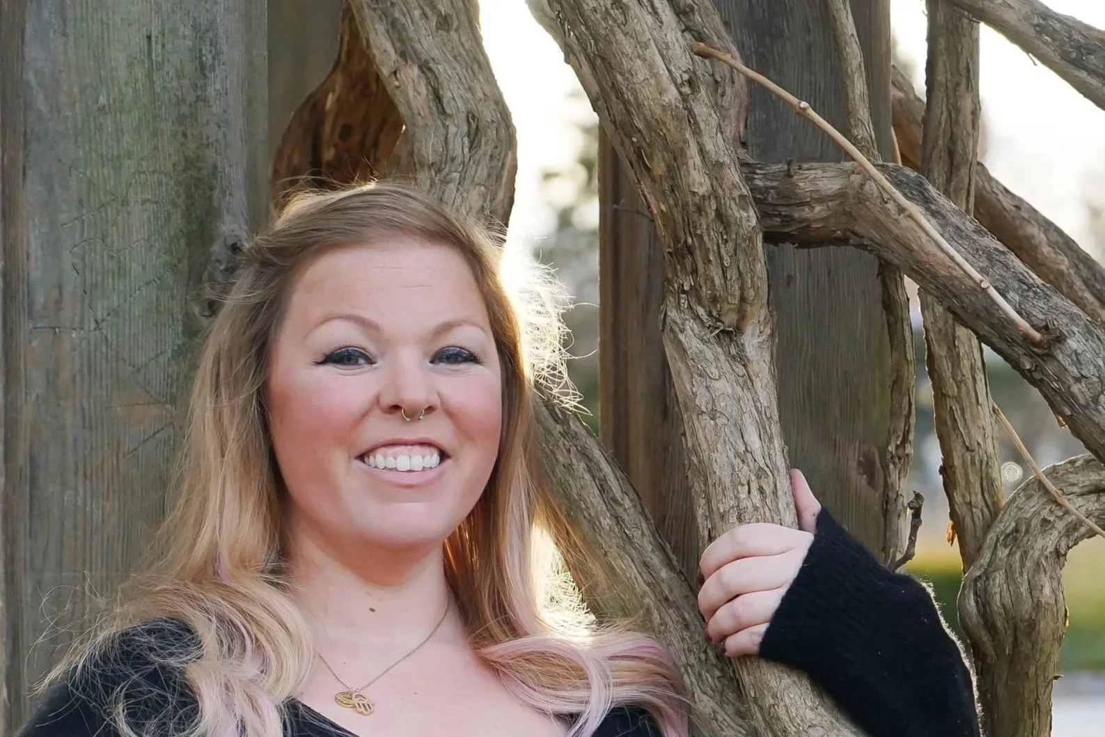 A smiling woman with reddish-blonde hair, a septum piercing, and a necklace, standing outdoors near a tree with twisted branches.