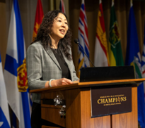 Woman speaking at a podium with flags in the background.