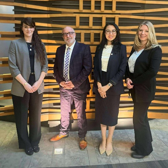 Four professionally dressed individuals in front of a modern wood backdrop.